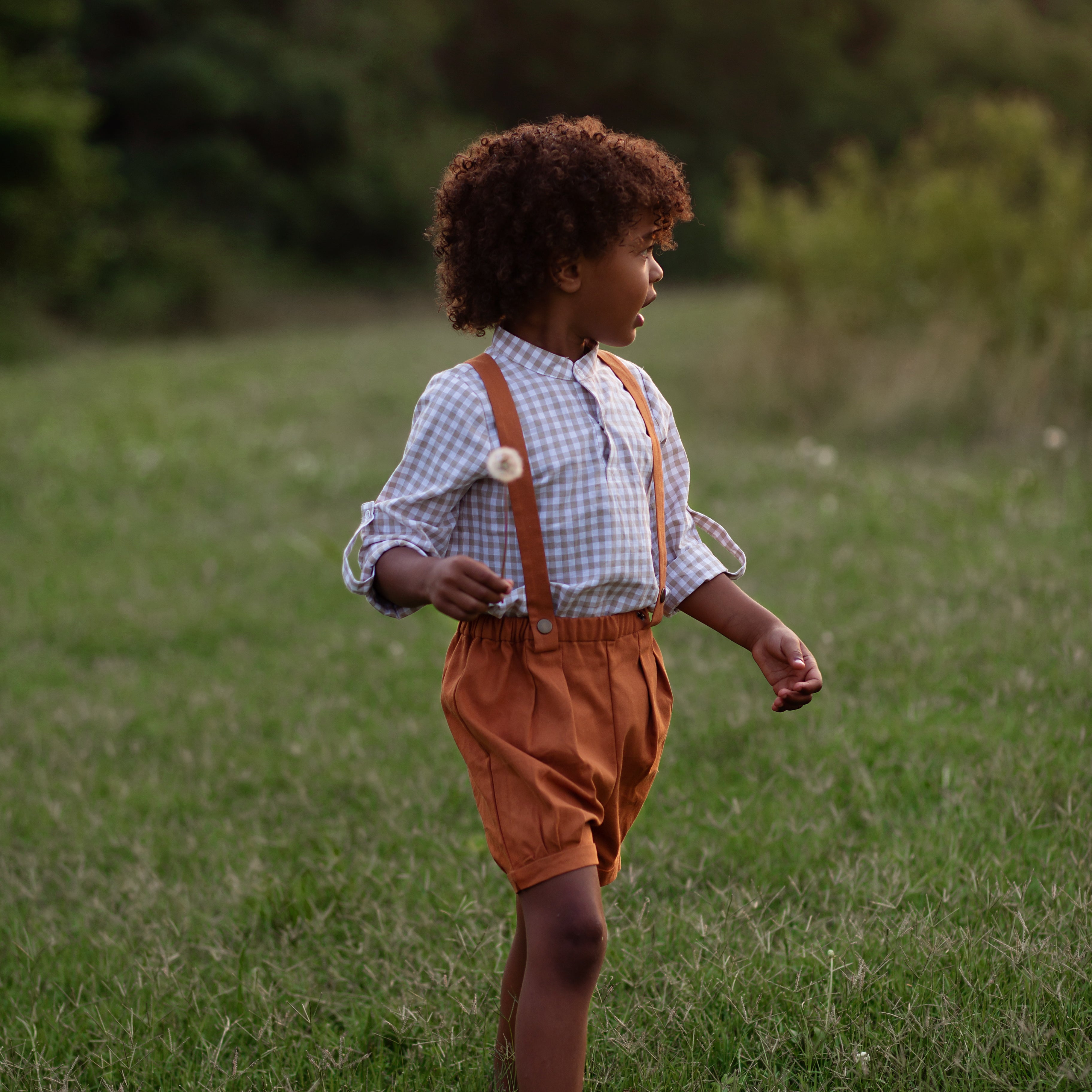 Model boy wearing the Burnt Orange Suspender Shorts with Beige Gingham Henley Shirt, side view - This image captures the side profile, showcasing the adjustable straps and overall silhouette of the outfit