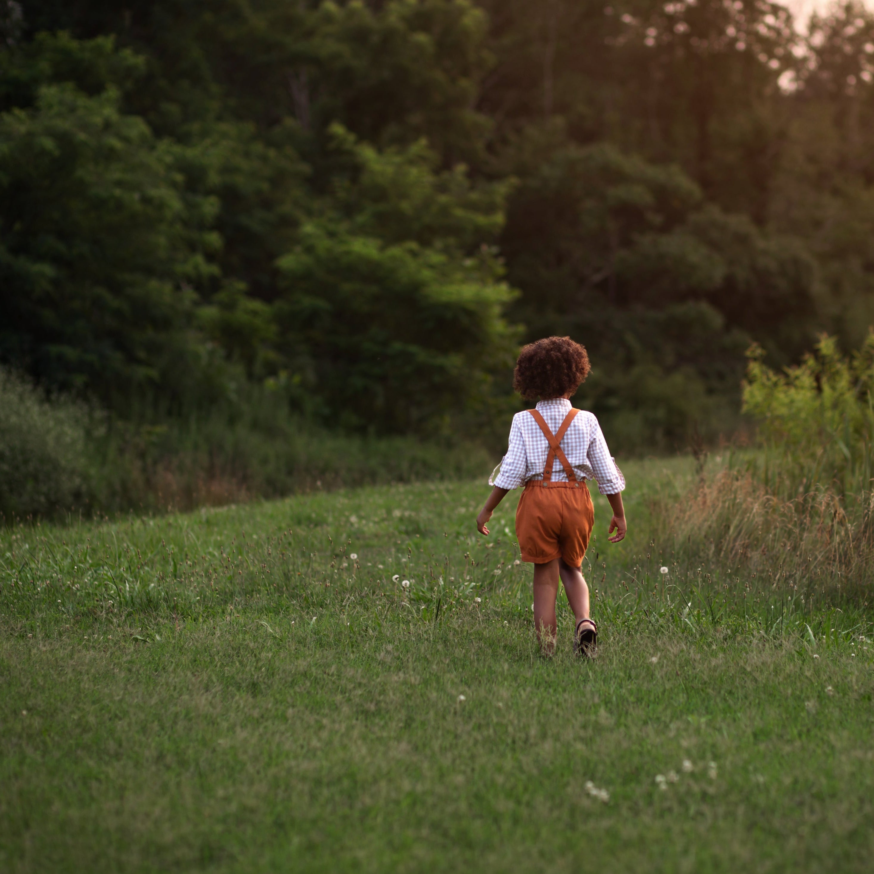 Back view of Beige Gingham Henley Shirt worn by 5-year-old boy in size 5Y, featuring a classic Henley placket and 3/4 cuffed sleeves that can be unrolled for layering. Paired with burnt orange linen suspender shorts for a stylish, versatile outfit.