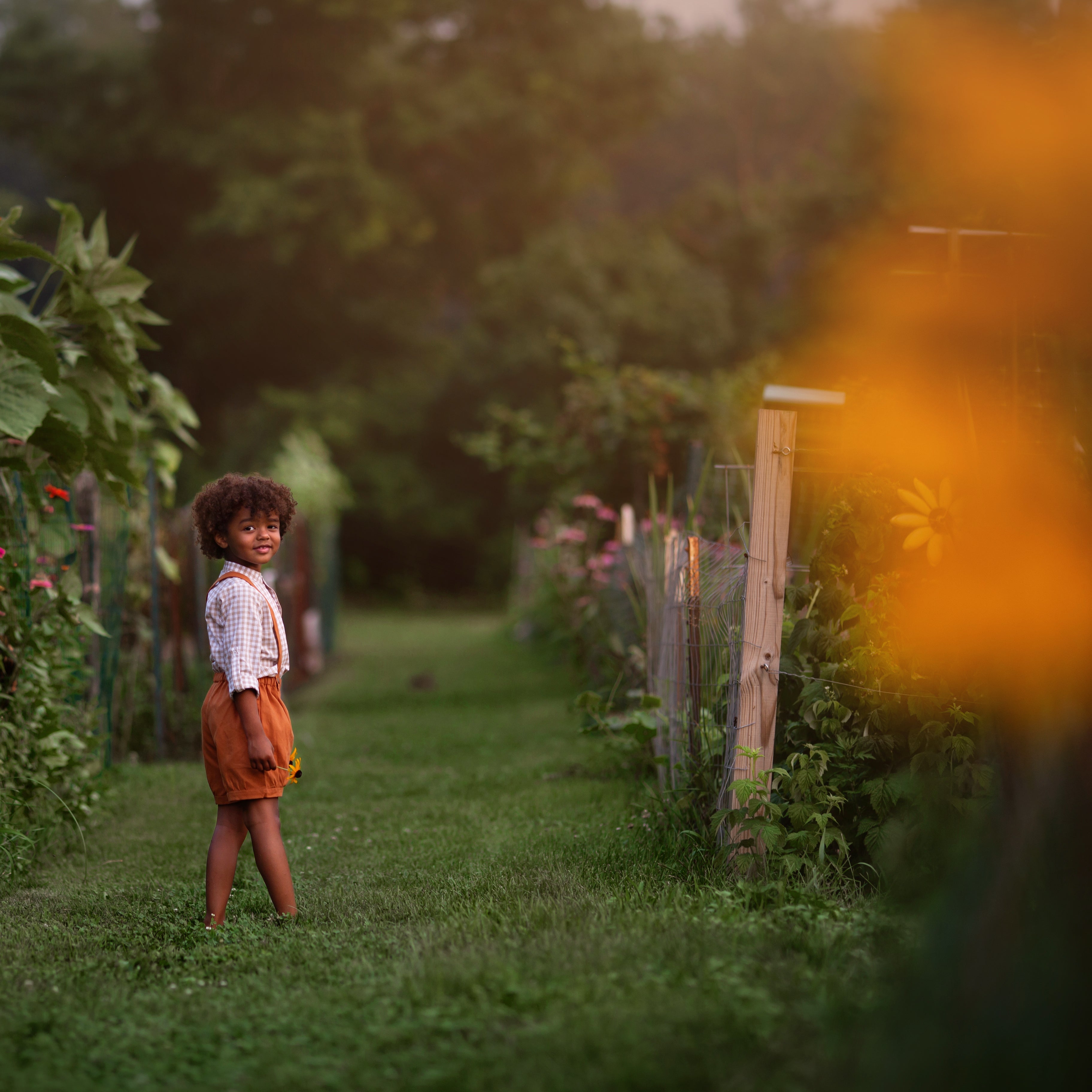 Model boy wearing the Burnt Orange Suspender Shorts with Beige Gingham Henley Shirt, far away view - A wider shot of the model, allowing for a full view of the outfit in a playful setting, emphasizing the versatility and style.