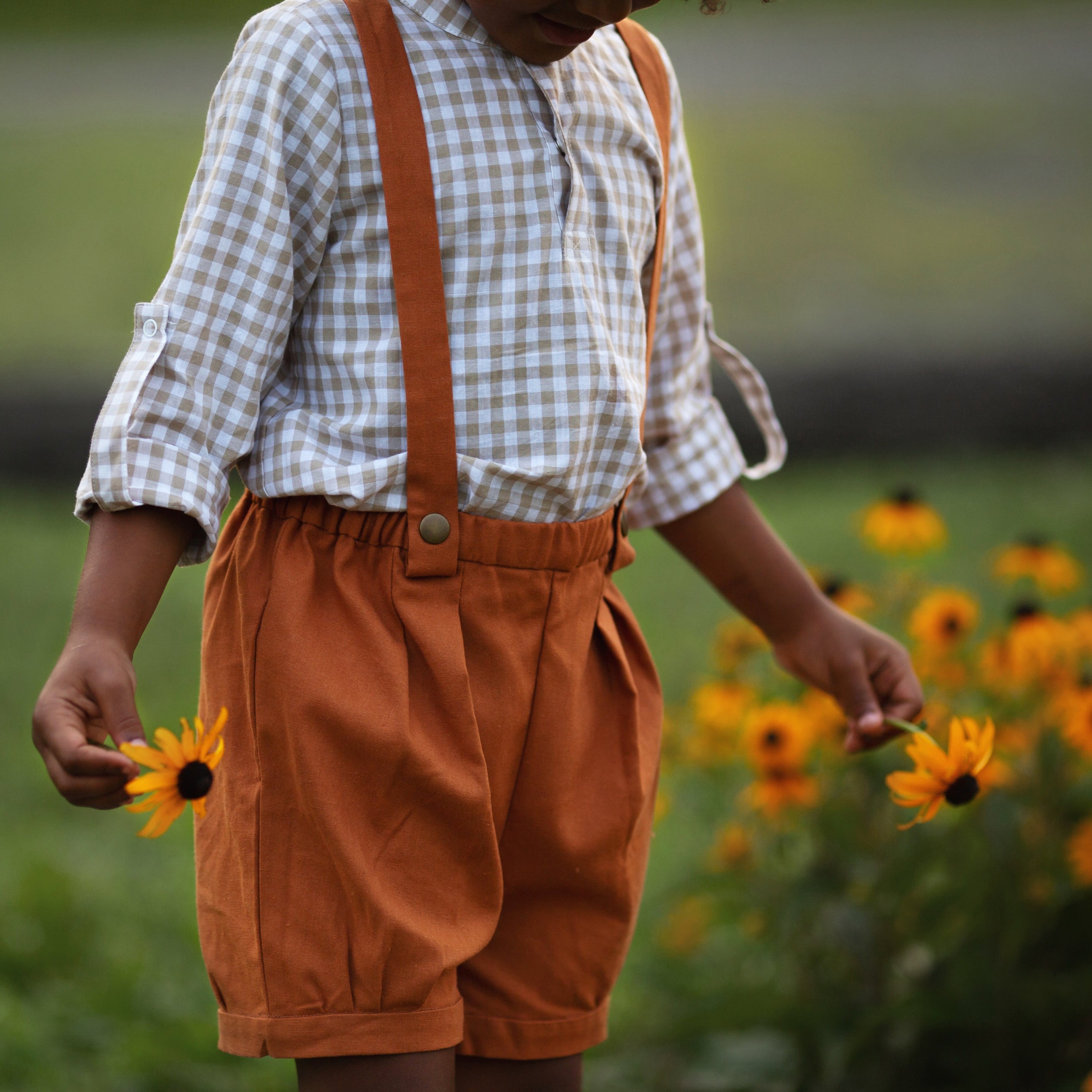 Detail of a beige Gingham Henley Shirt worn by 5-year-old boy in size 5Y, featuring a classic Henley placket and 3/4 cuffed sleeves that can be unrolled for layering. Paired with burnt orange linen suspender shorts for a stylish, versatile outfit.