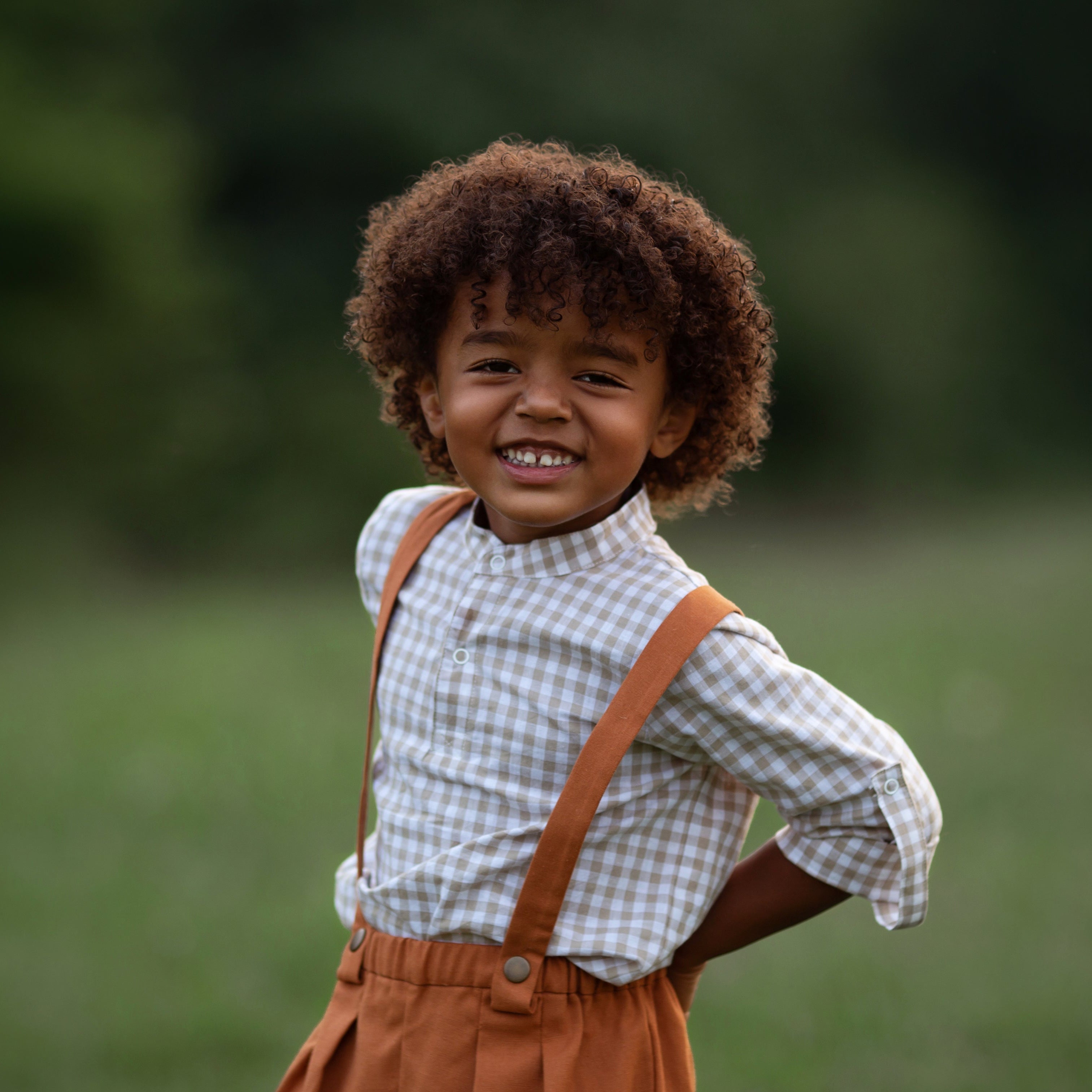 Beige Gingham Henley Shirt worn by a laughing 5-year-old boy in size 5Y, featuring a classic Henley placket and 3/4 cuffed sleeves that can be unrolled for layering. Paired with burnt orange linen suspender shorts for a stylish, versatile outfit.
