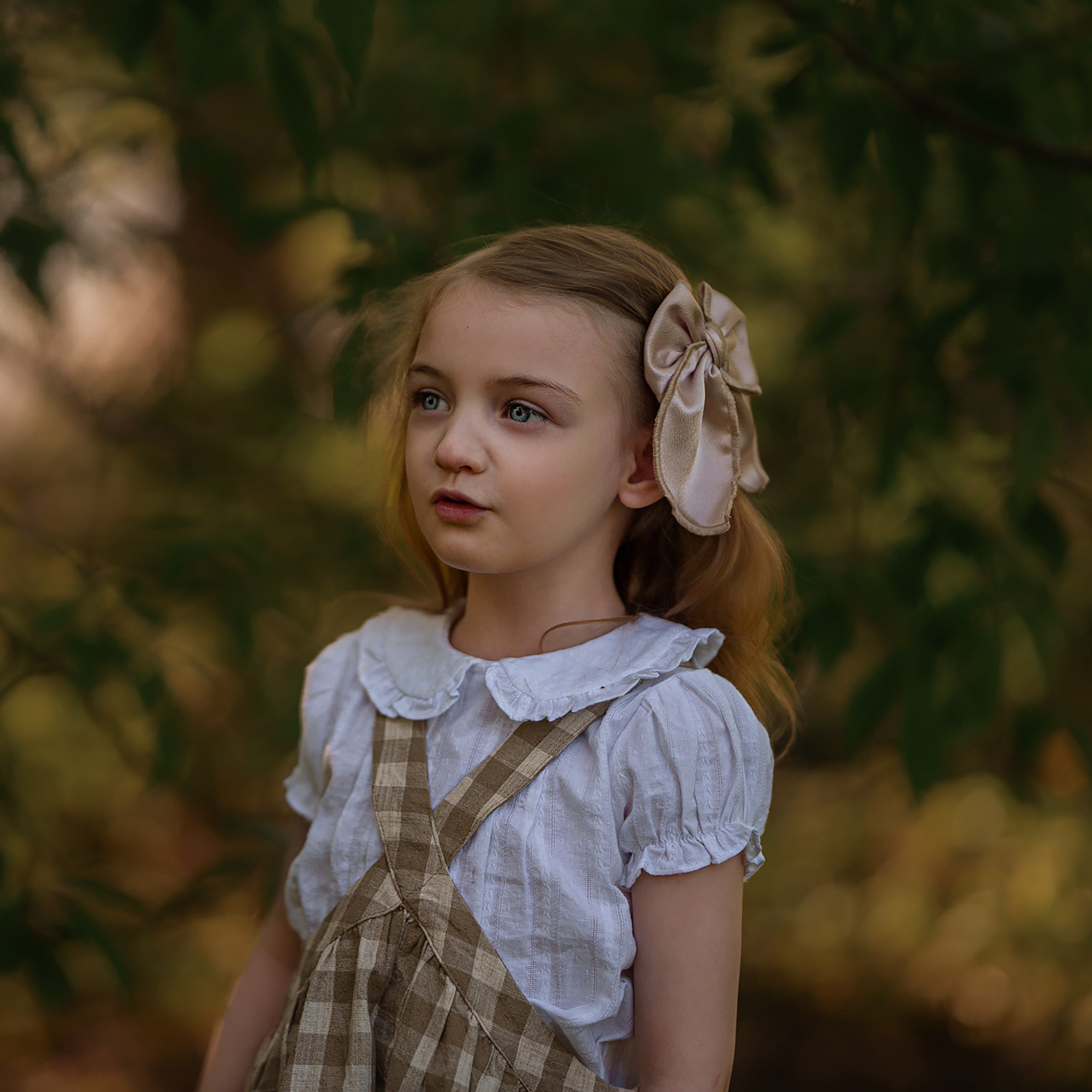 Young girl in a white blouse and checkered dress with a bow in her hair, standing in a natural setting.