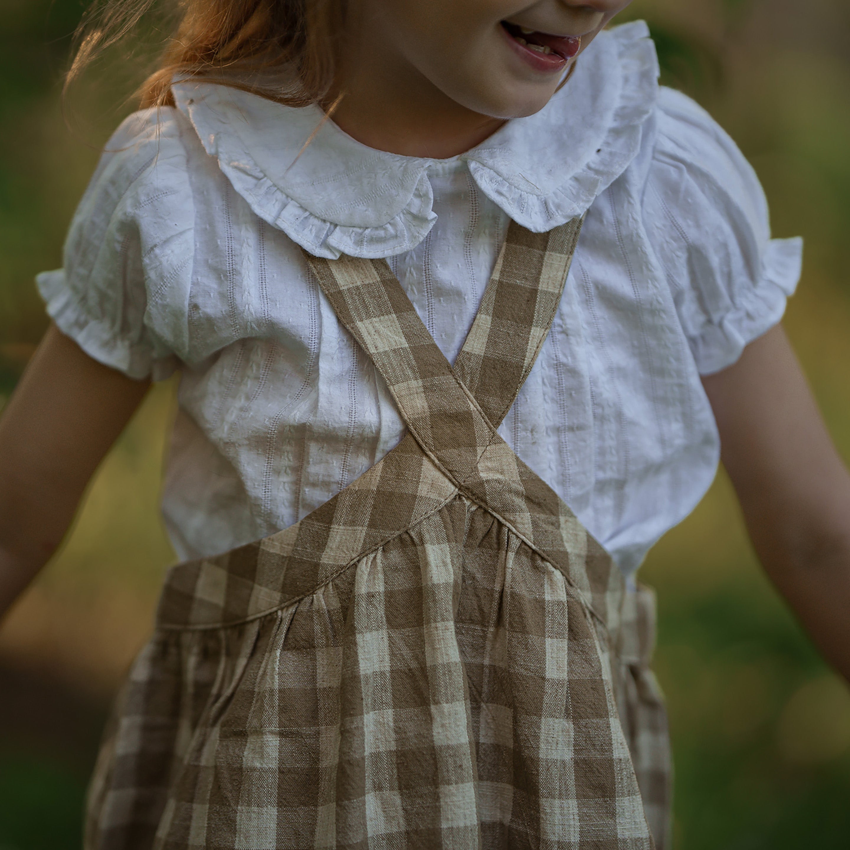 Child wearing a white blouse with ruffled collar and brown checkered overalls outdoors.