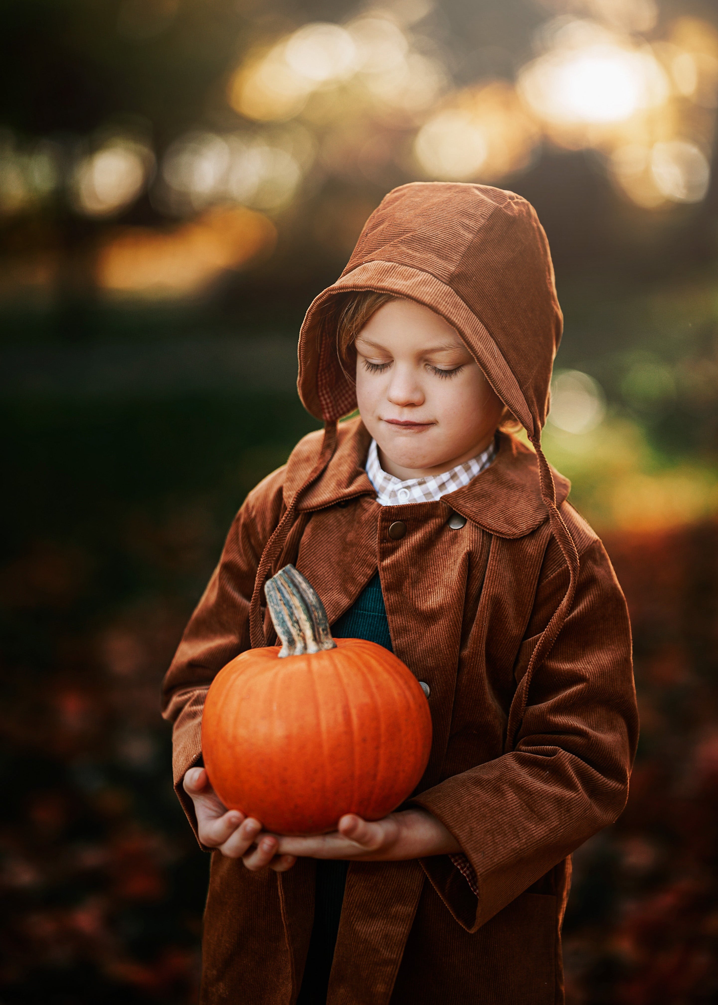 Child in a brown coat holding a small pumpkin with a blurred outdoor background