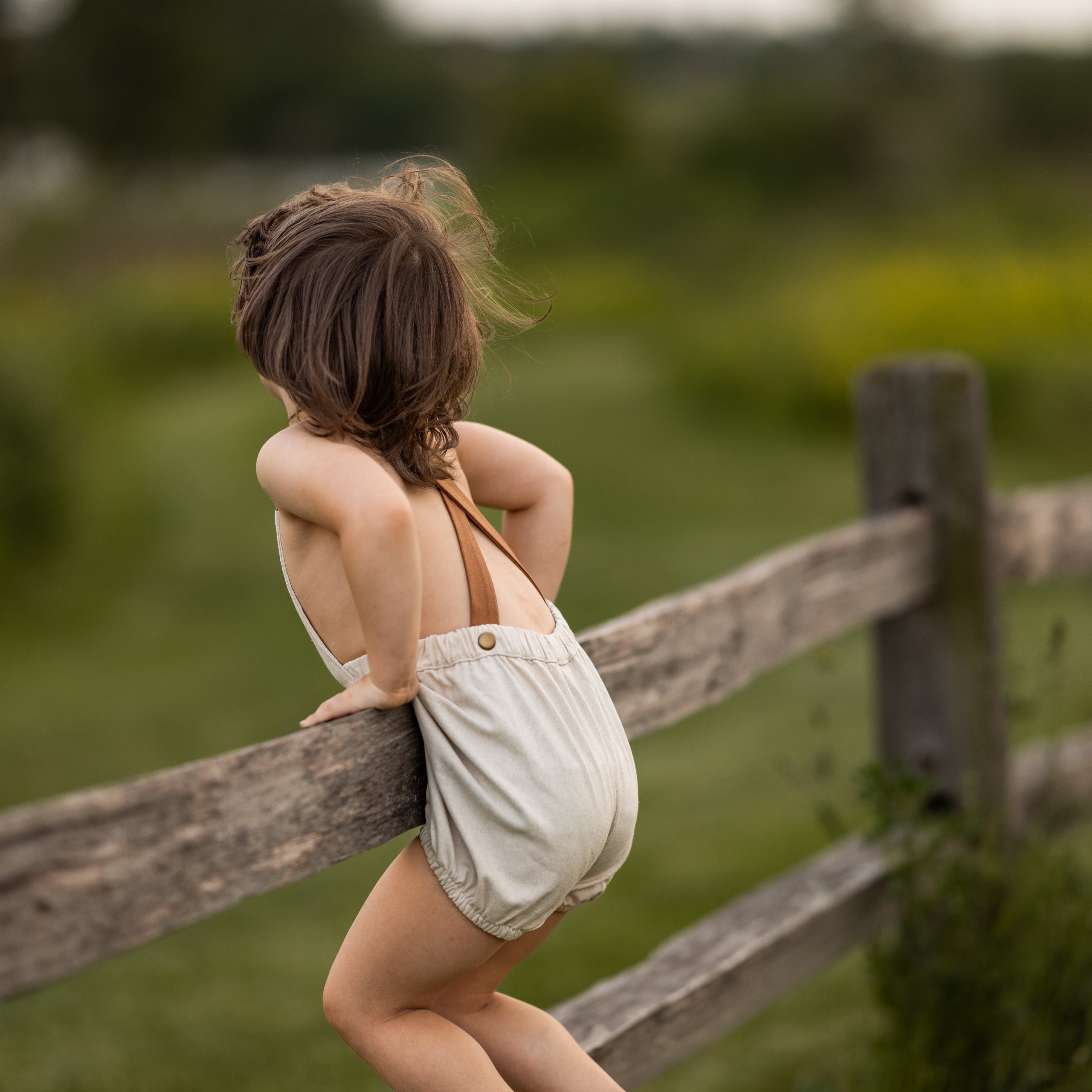 Back view of a Toddler boy wearing a handmade linen bubble romper with contrasting straps, playing in a lush green field. The romper is designed for comfort and style, perfect for outdoor summer activities.