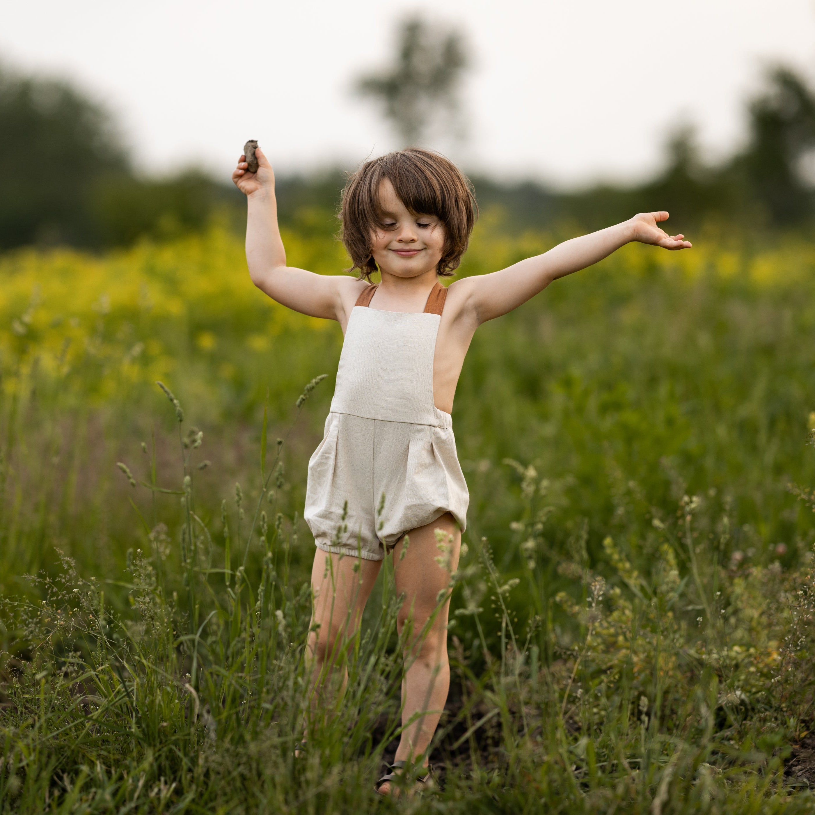 Toddler boy wearing a handmade linen bubble romper with contrasting straps, playing in a lush green field. The romper is designed for comfort and style, perfect for outdoor summer activities.