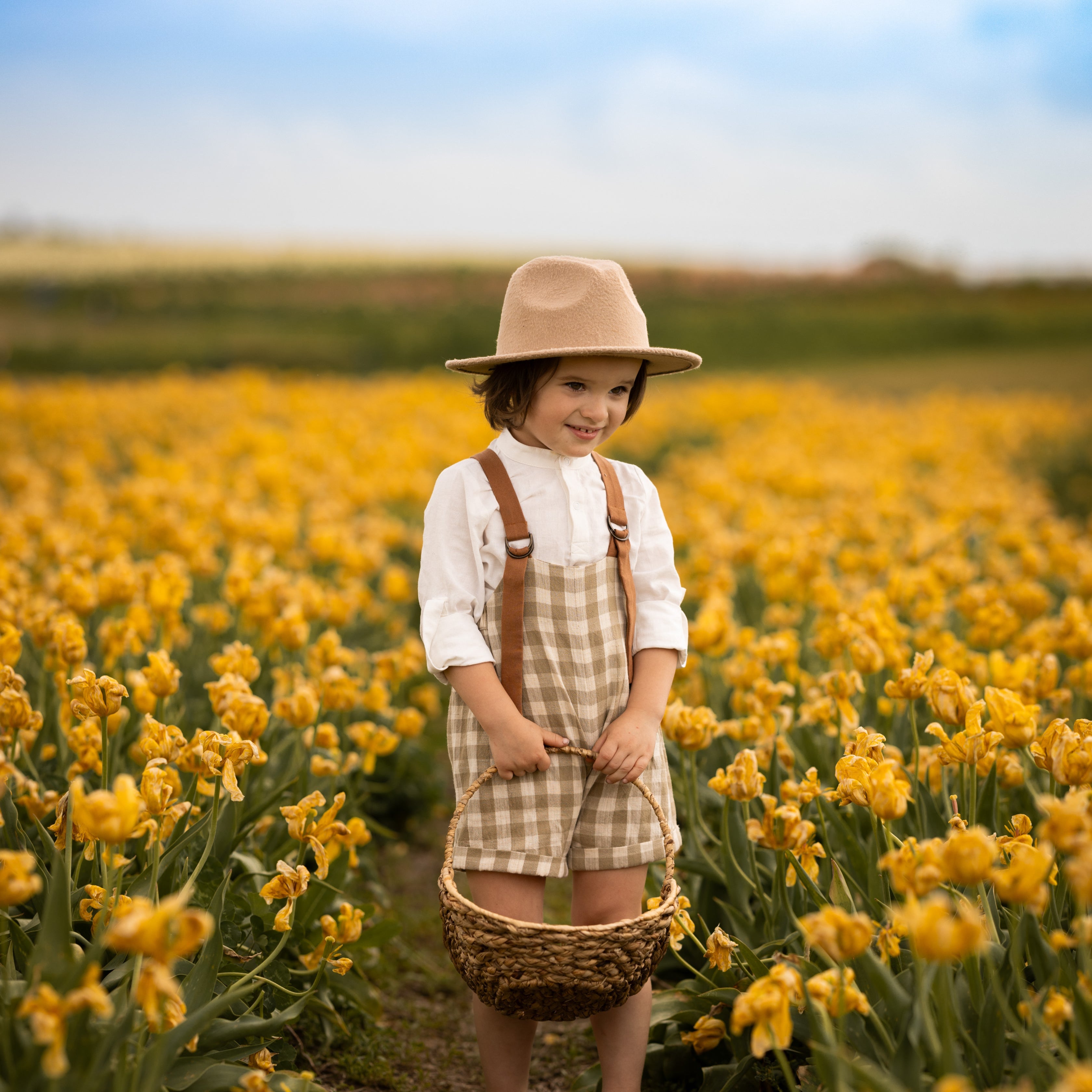 Child in a hat and checkered dress standing in a field of yellow flowers holding a basket.