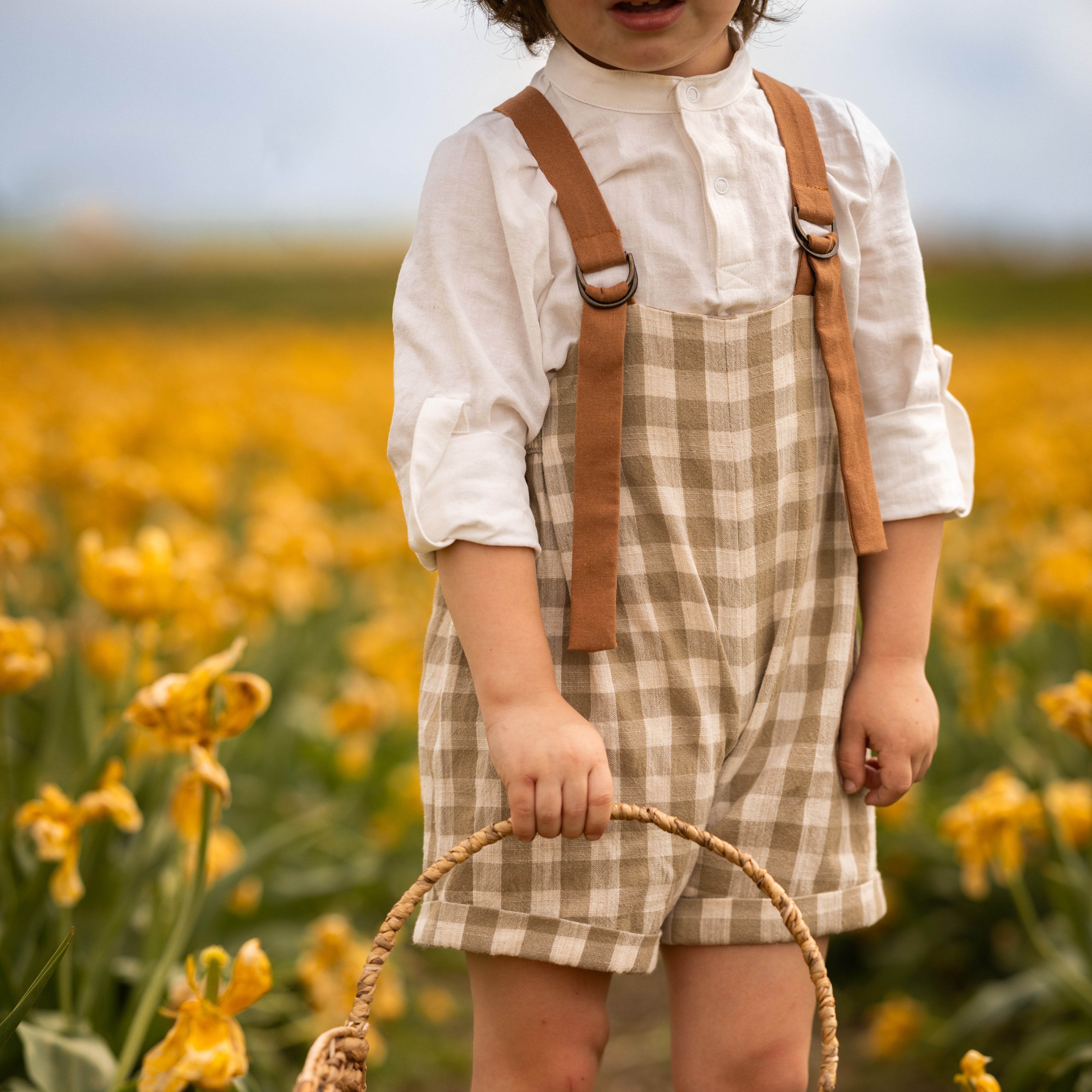 Little boy in a beige gingham romper surrounded by yellow flowers in a field.