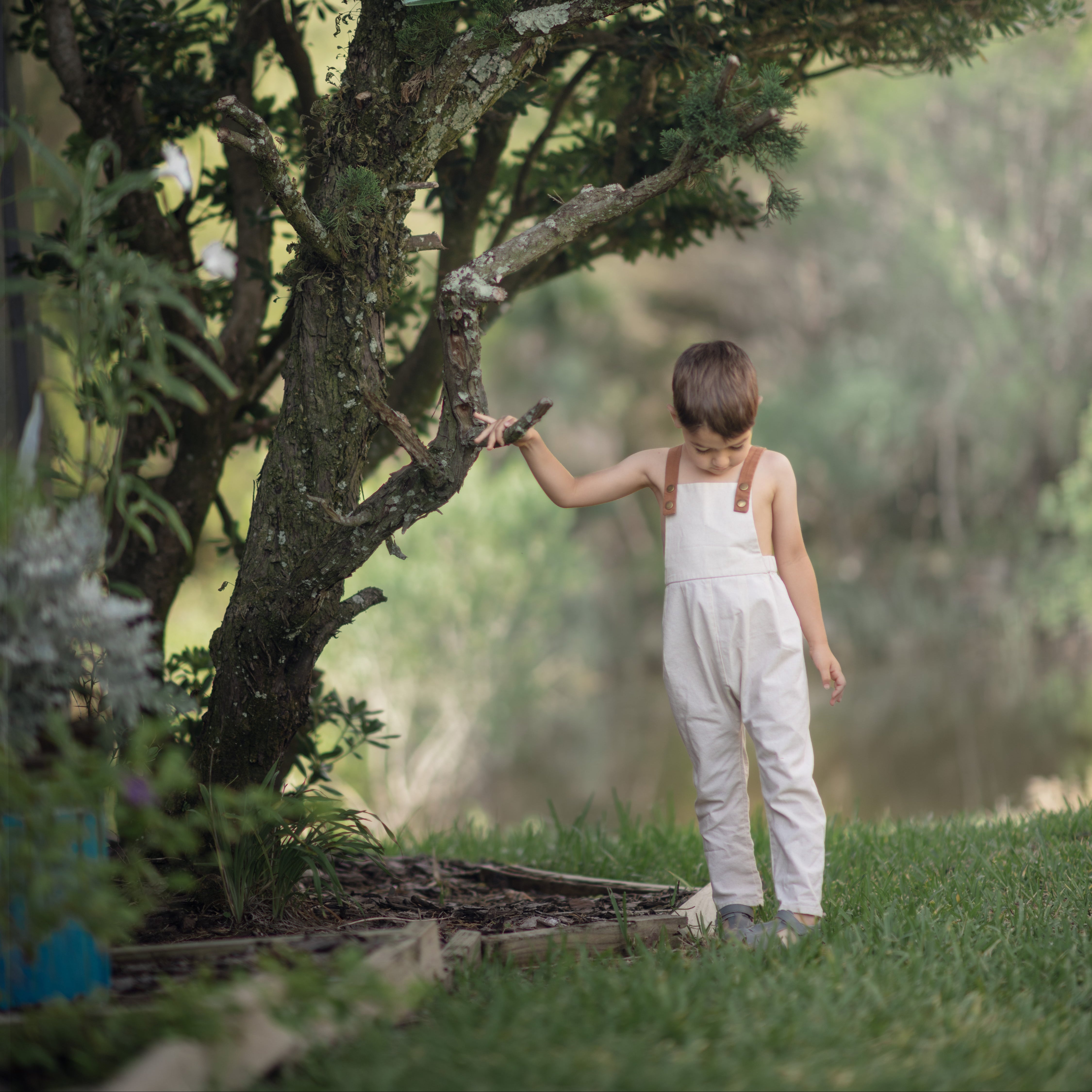 Child in natural linen overalls reaching towards a green watering can attached to a tree branch.
