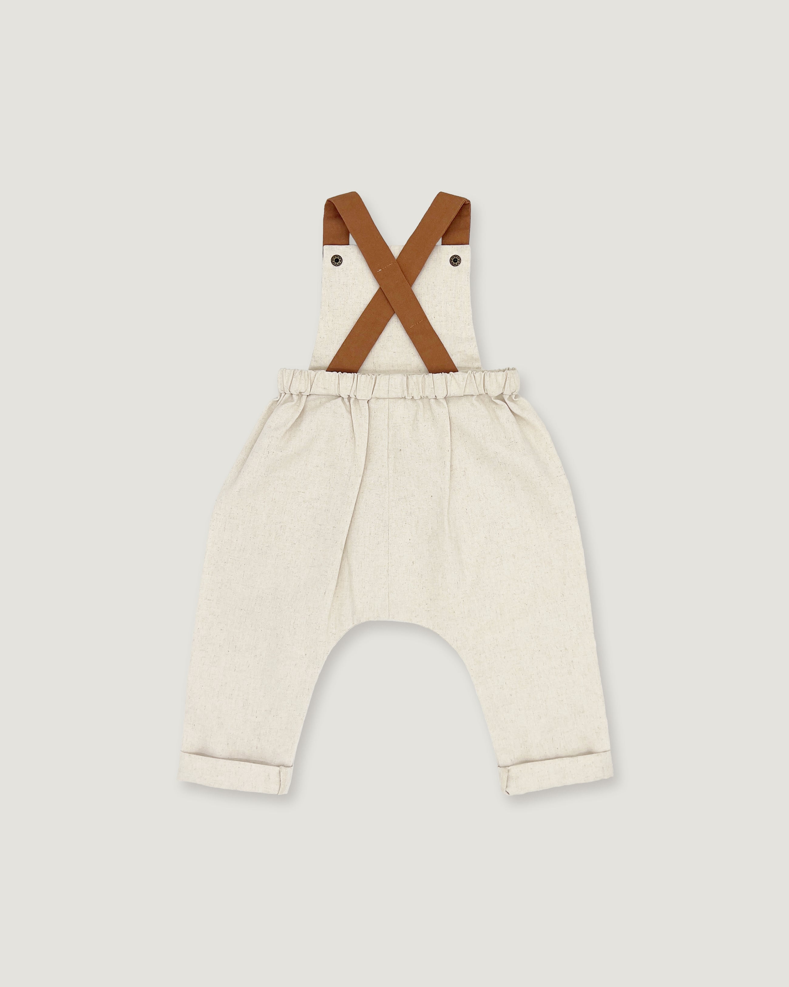 A pair of baby bib pants in a light cream linen-like fabric, featuring brown faux-leather suspenders that cross over the chest. The pants have an elastic waistband and rolled cuffs, shown against a solid light gray background.