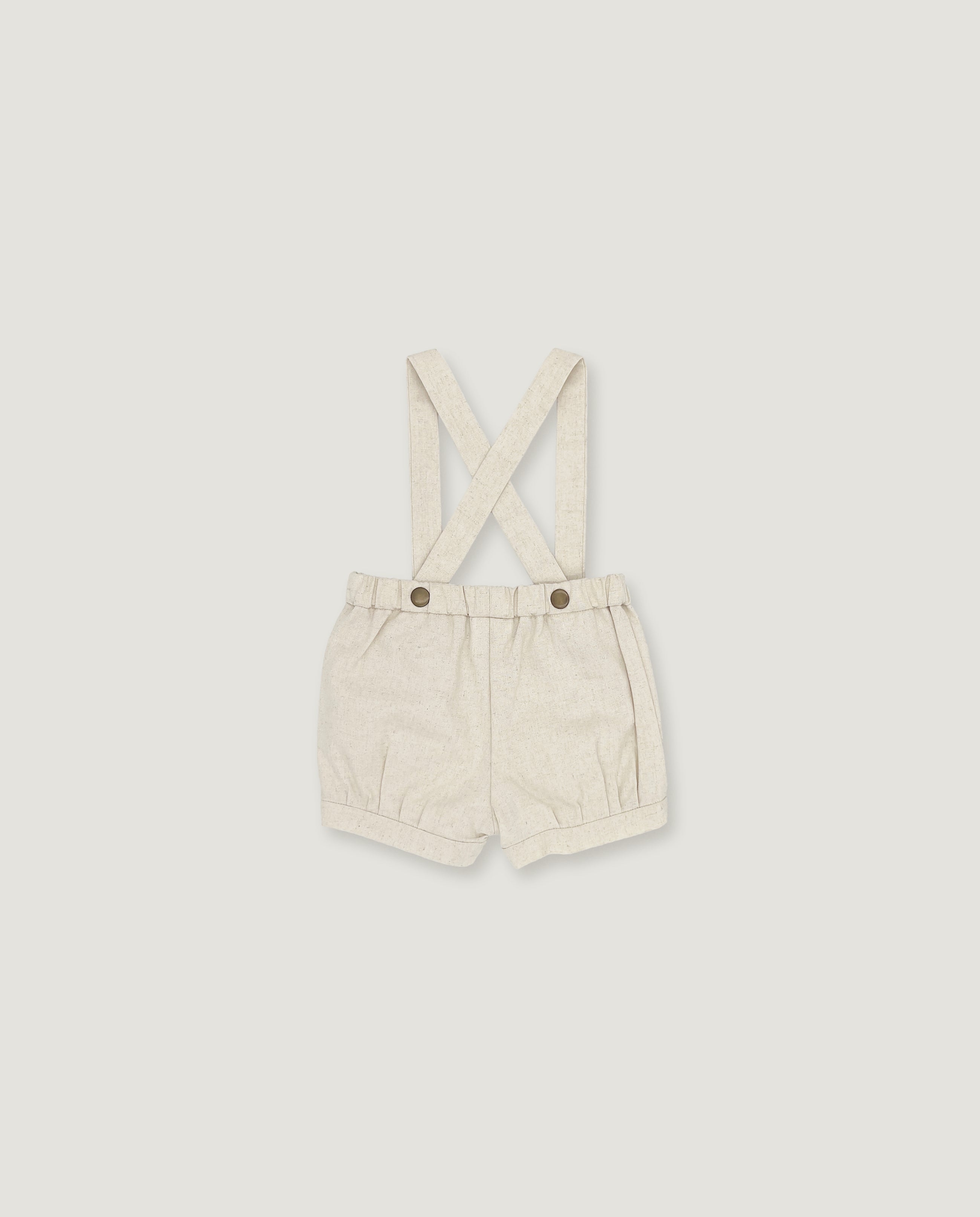 A pair of cream-colored linen baby shorts with adjustable shoulder straps and two metal snap buttons, displayed flat against a light gray background.