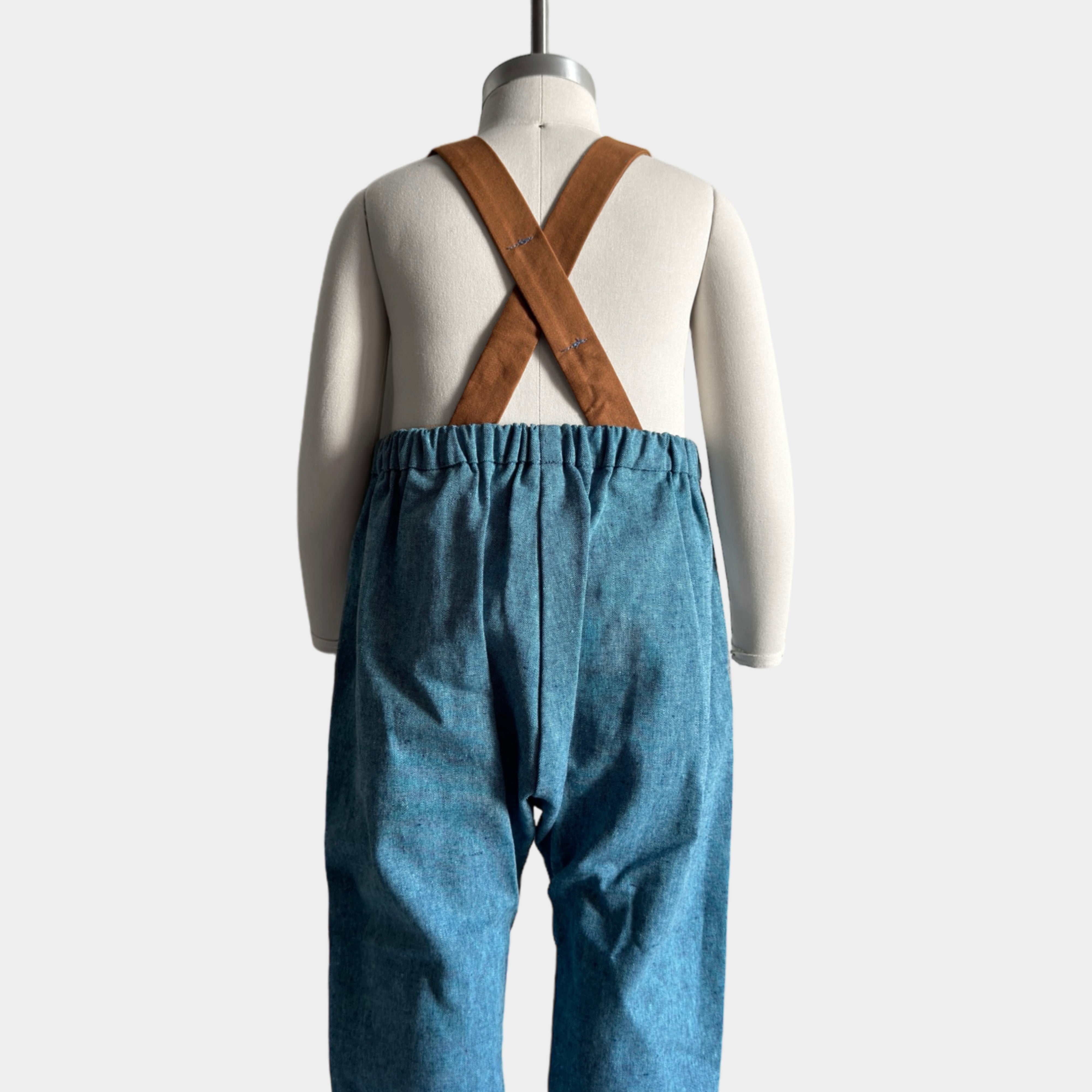 Blue Jay Long Overalls on a mannequin showcasing the back - Handmade from repurposed linen fabric, these gender-neutral overalls feature visually appealing brown leather-looking straps that enhance durability. The breathable fabric ensures all-day comfort, while the stylish pleats add elegance. Adjustable straps in two lengths provide a perfect fit, and the unfoldable leg cuffs are designed to grow with your little one, making these overalls a practical and sustainable choice.