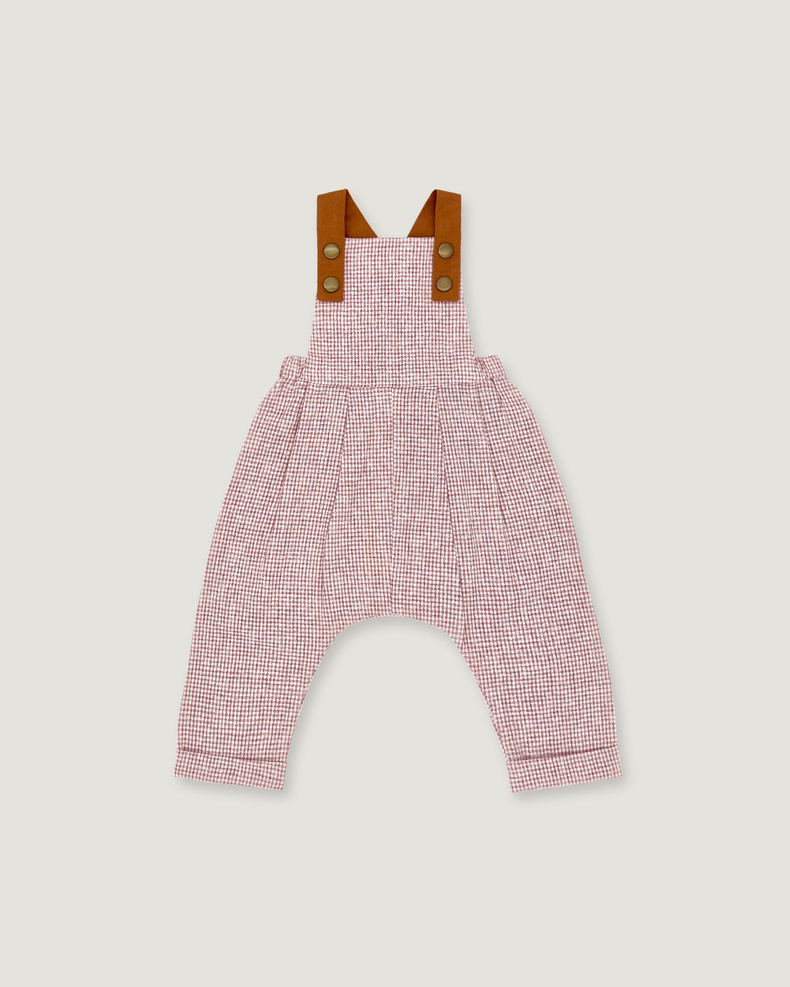 A baby romper with a pink and white micro-check pattern, featuring brown leather-like shoulder straps with snap buttons, shown flat on a light gray background.