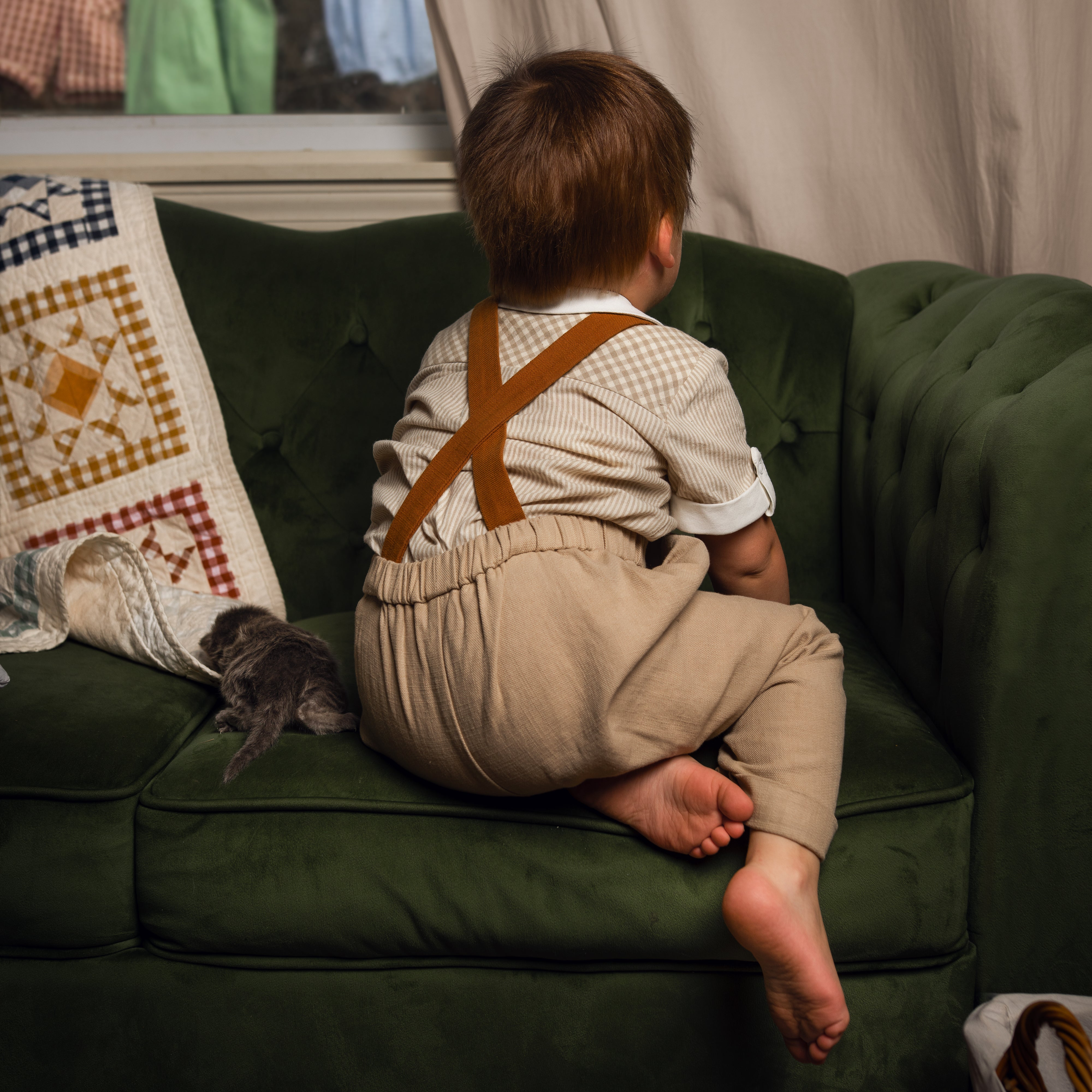 Child in suspenders sitting on a green couch with a cat nearby