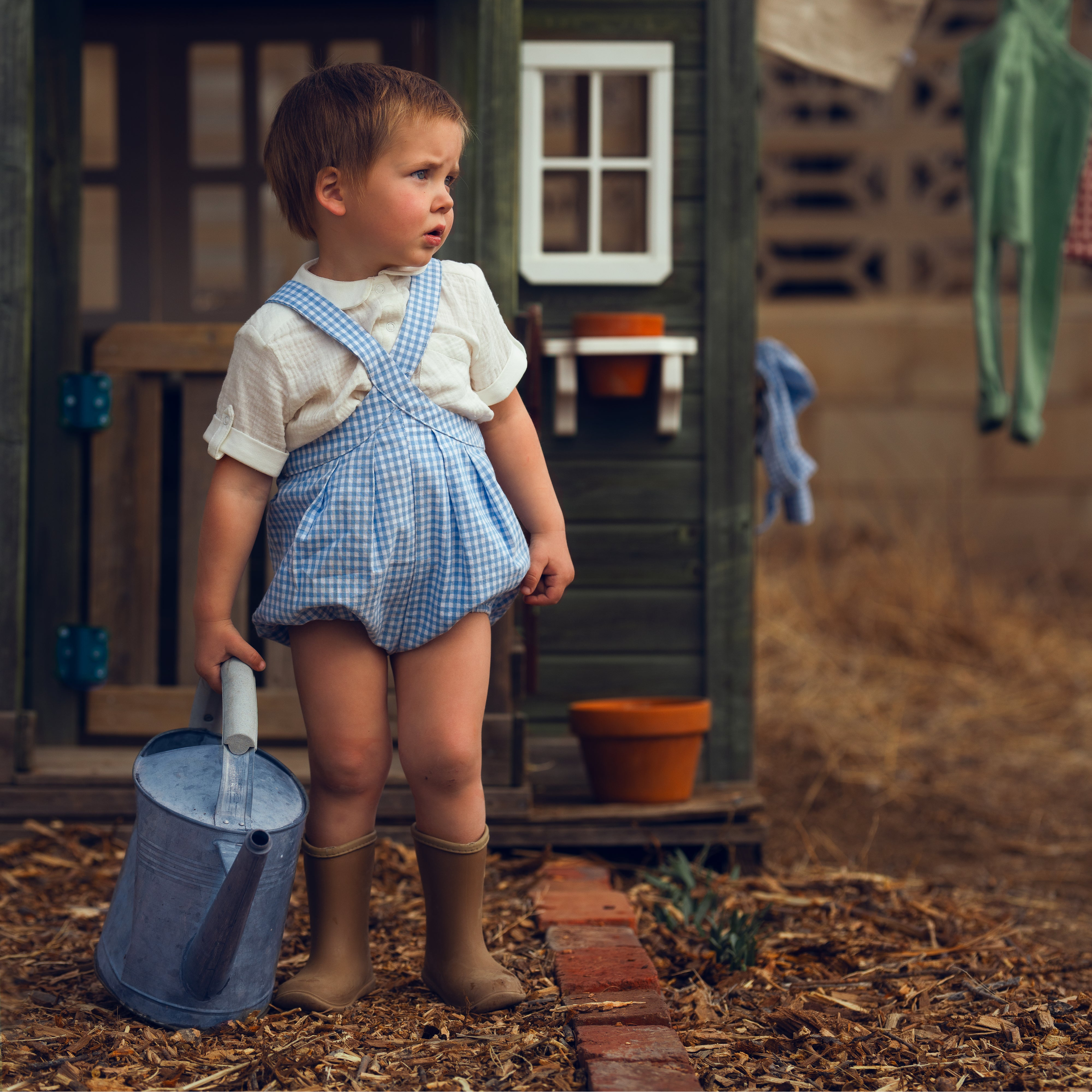 Child in checkered overalls standing in front of a wooden playhouse