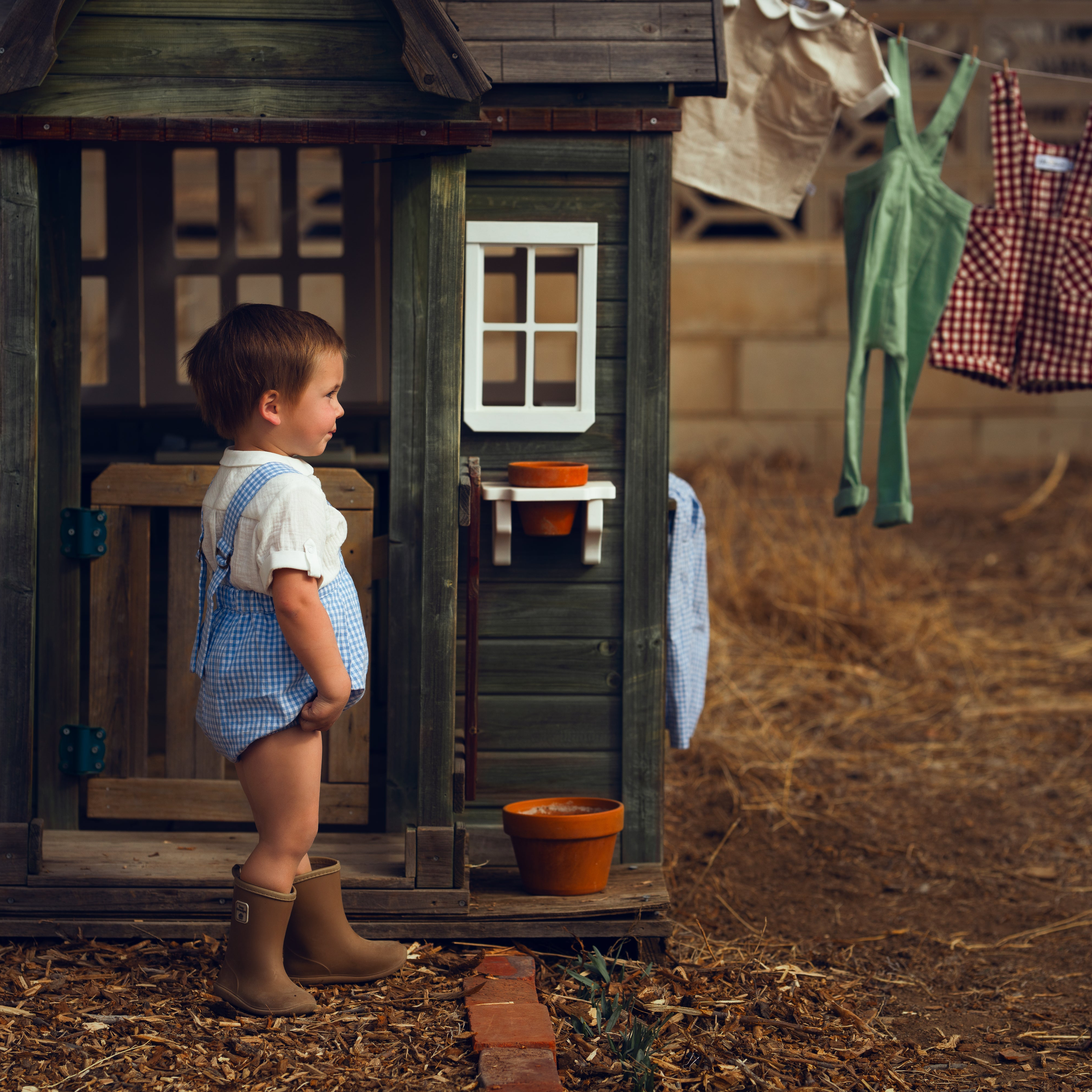 Child standing in front of a wooden playhouse with clothes hanging on a line in the background.