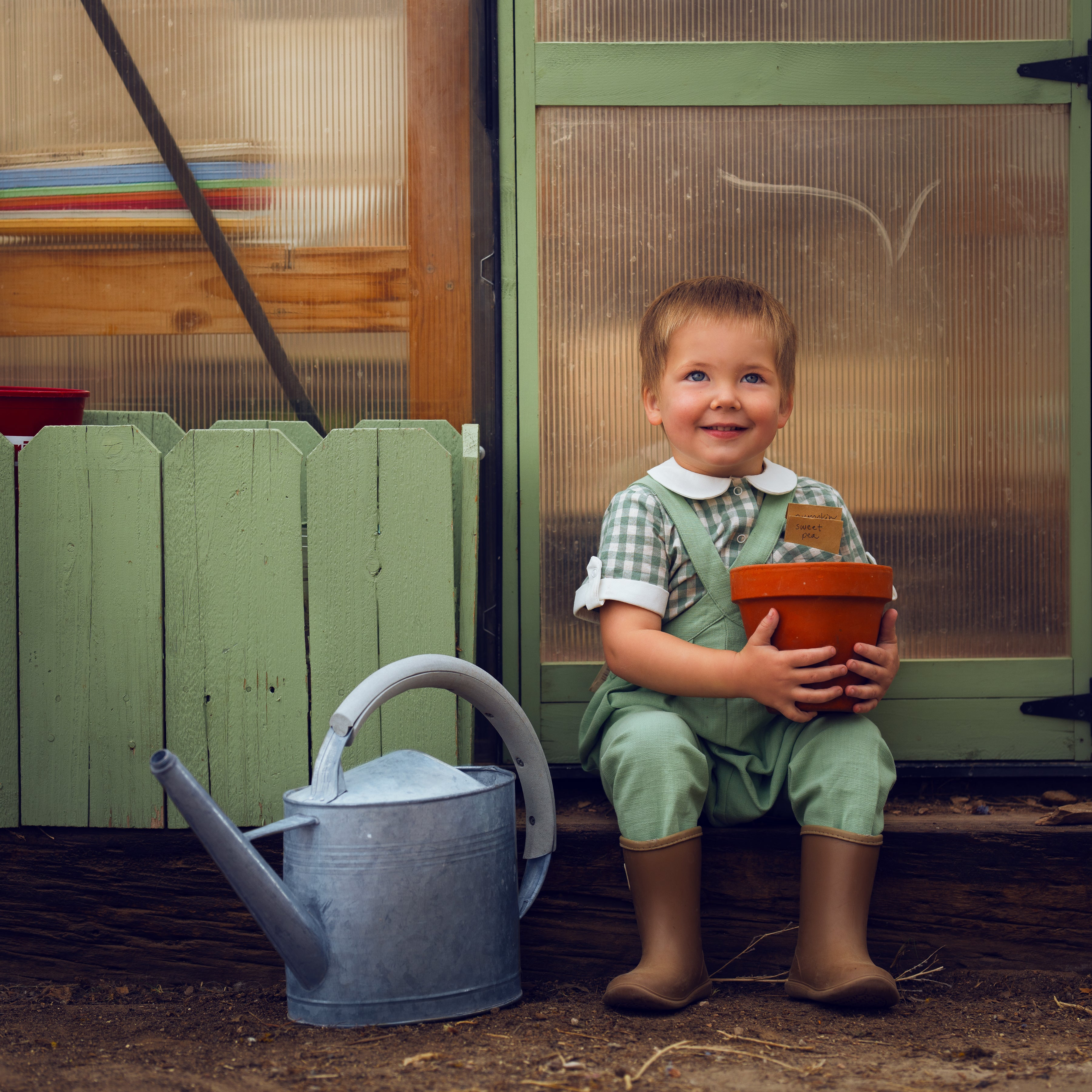 Child holding a pot and smiling in front of a green door with a watering can on the ground.