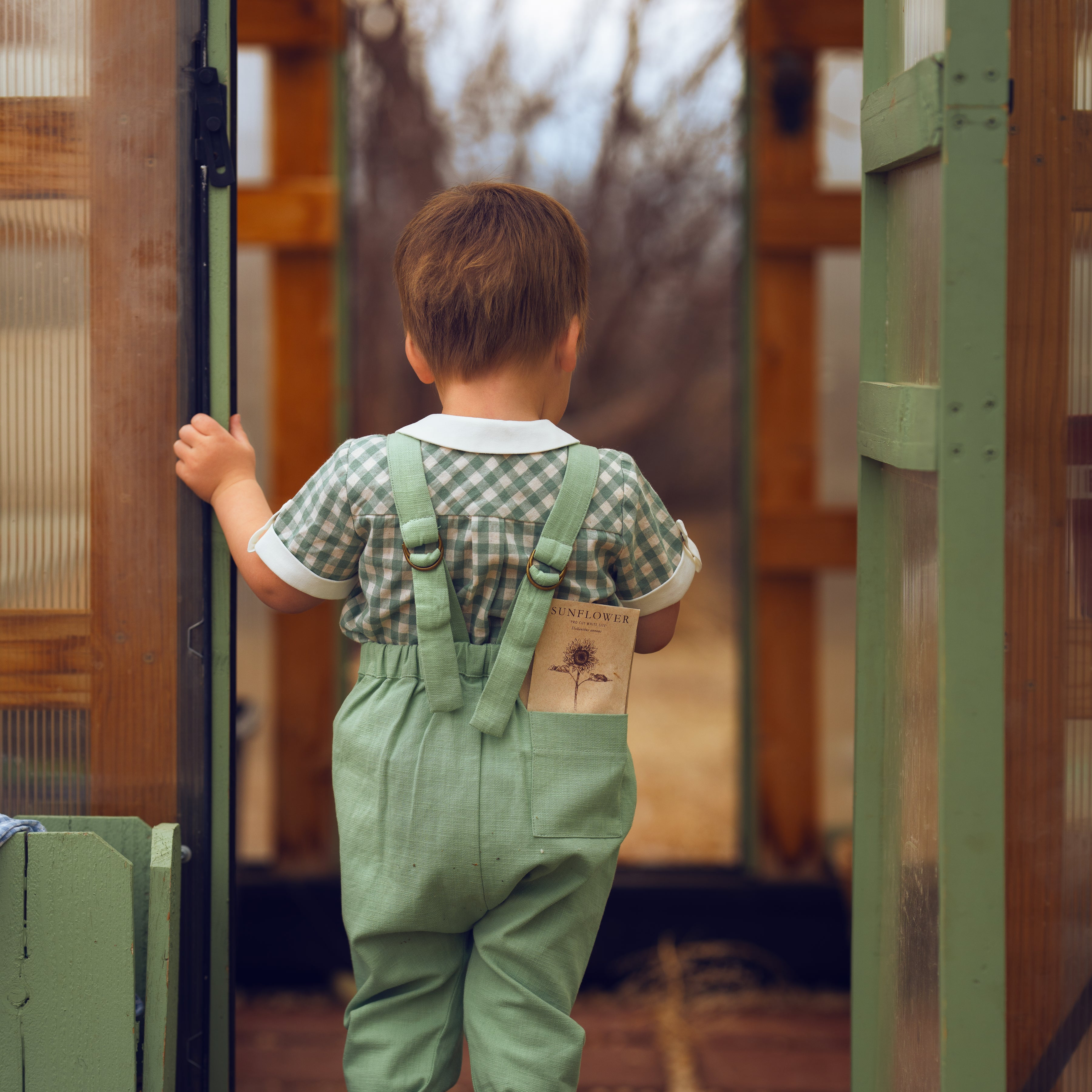 Child wearing green overalls and brown boots standing inside a wooden shed.