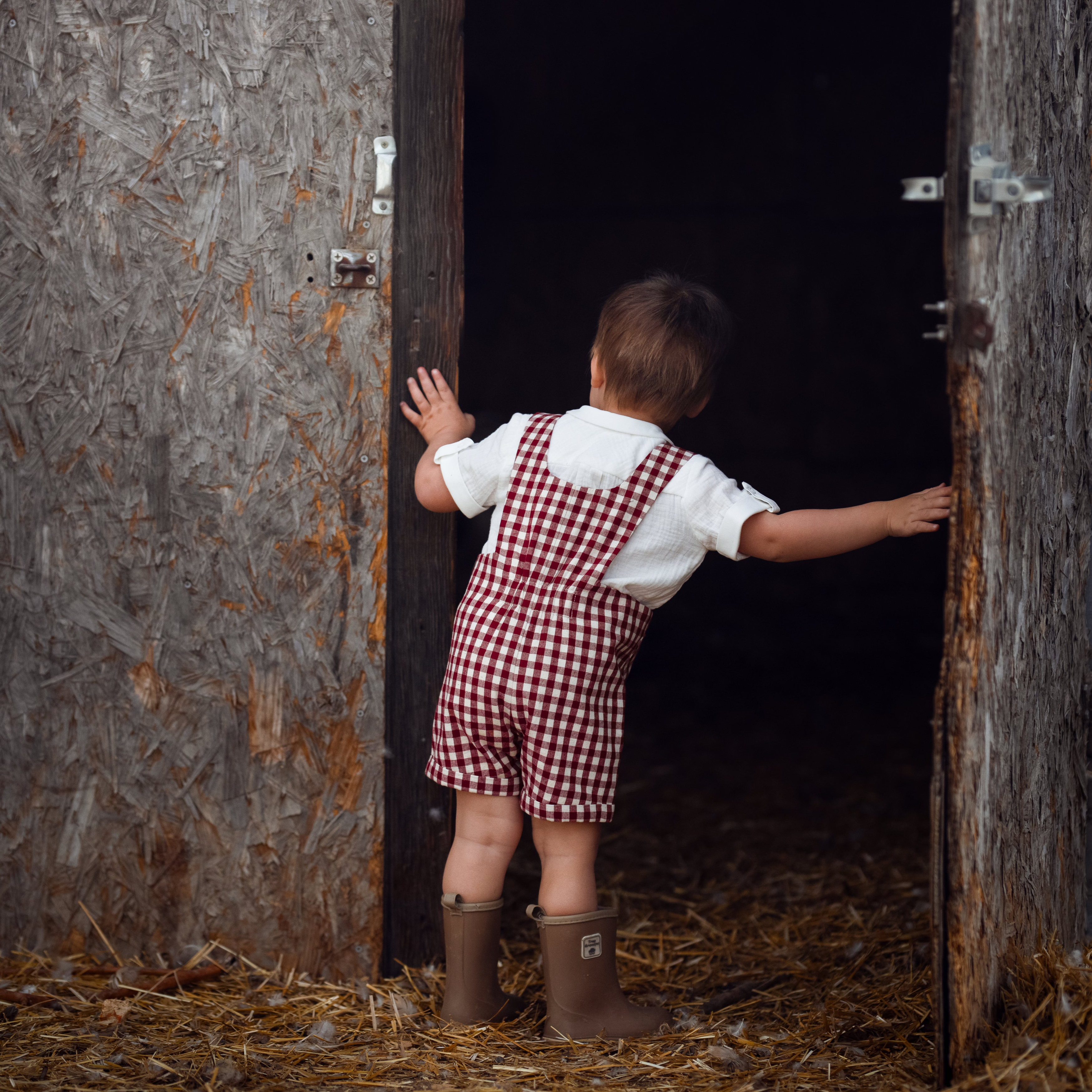 Child in checkered overalls and boots standing in a dark doorway with hay on the floor.