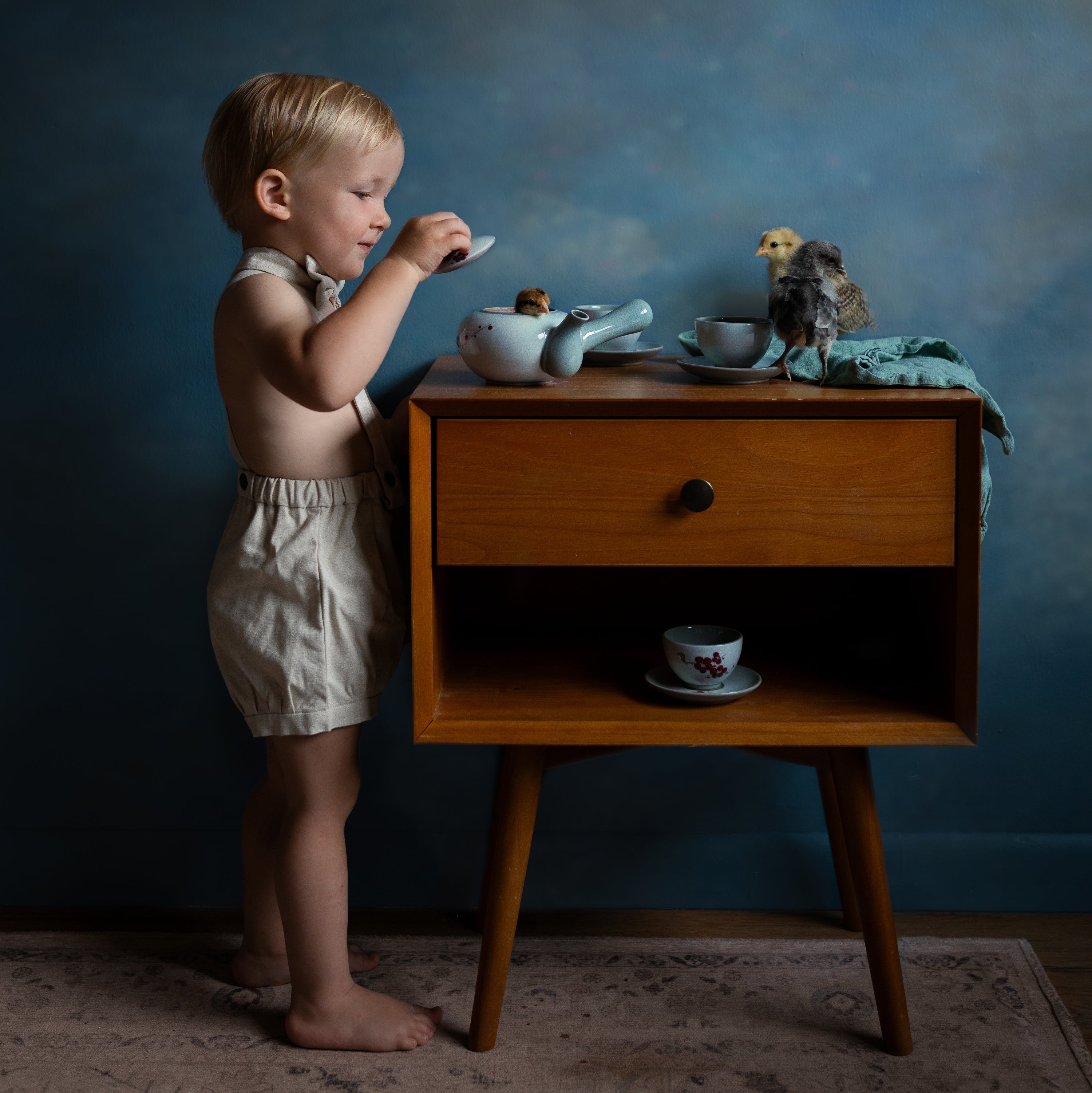 Boy in suspender shorts and matching boy tie playing with chicks on a piece of furniture