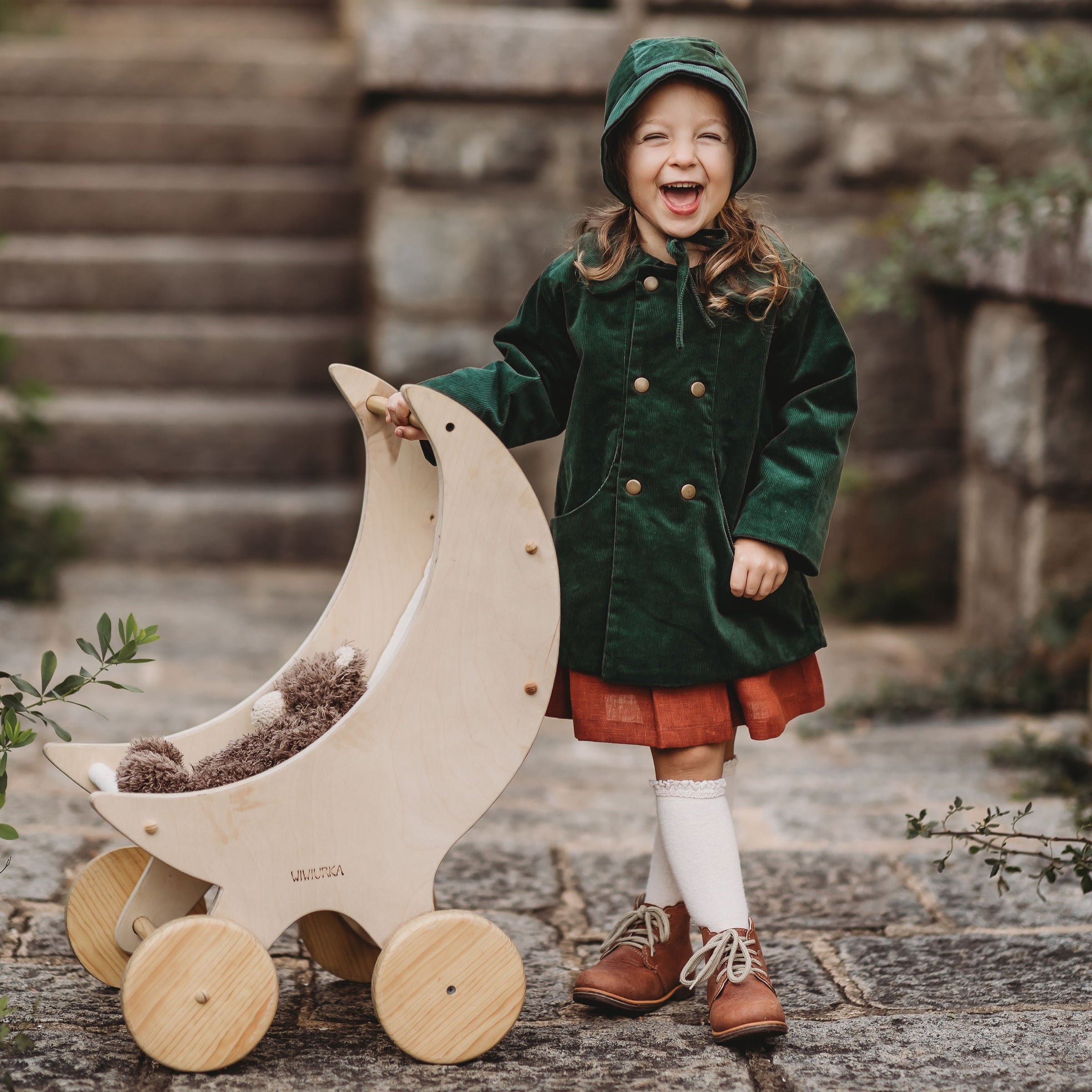 Child in a green coat and hat holding a wooden toy outdoors.