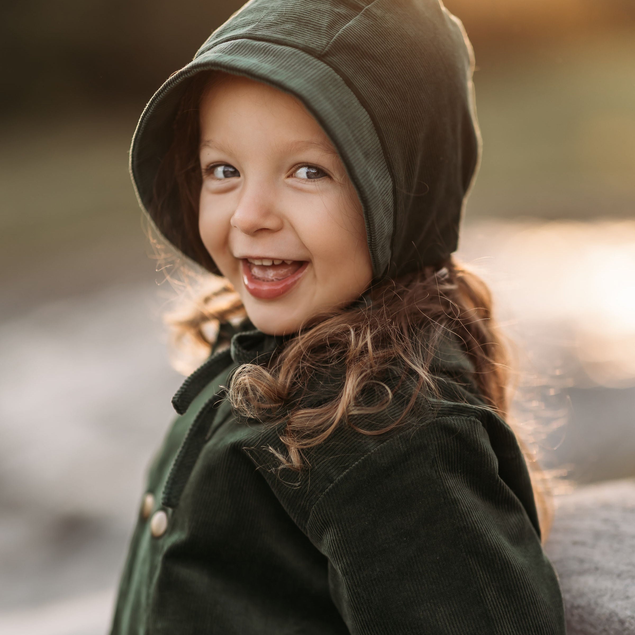 Smiling 3-year-old girl wearing the Evergreen corduroy bonnet and matching coat, highlighting the gender-neutral style and comfortable fit.