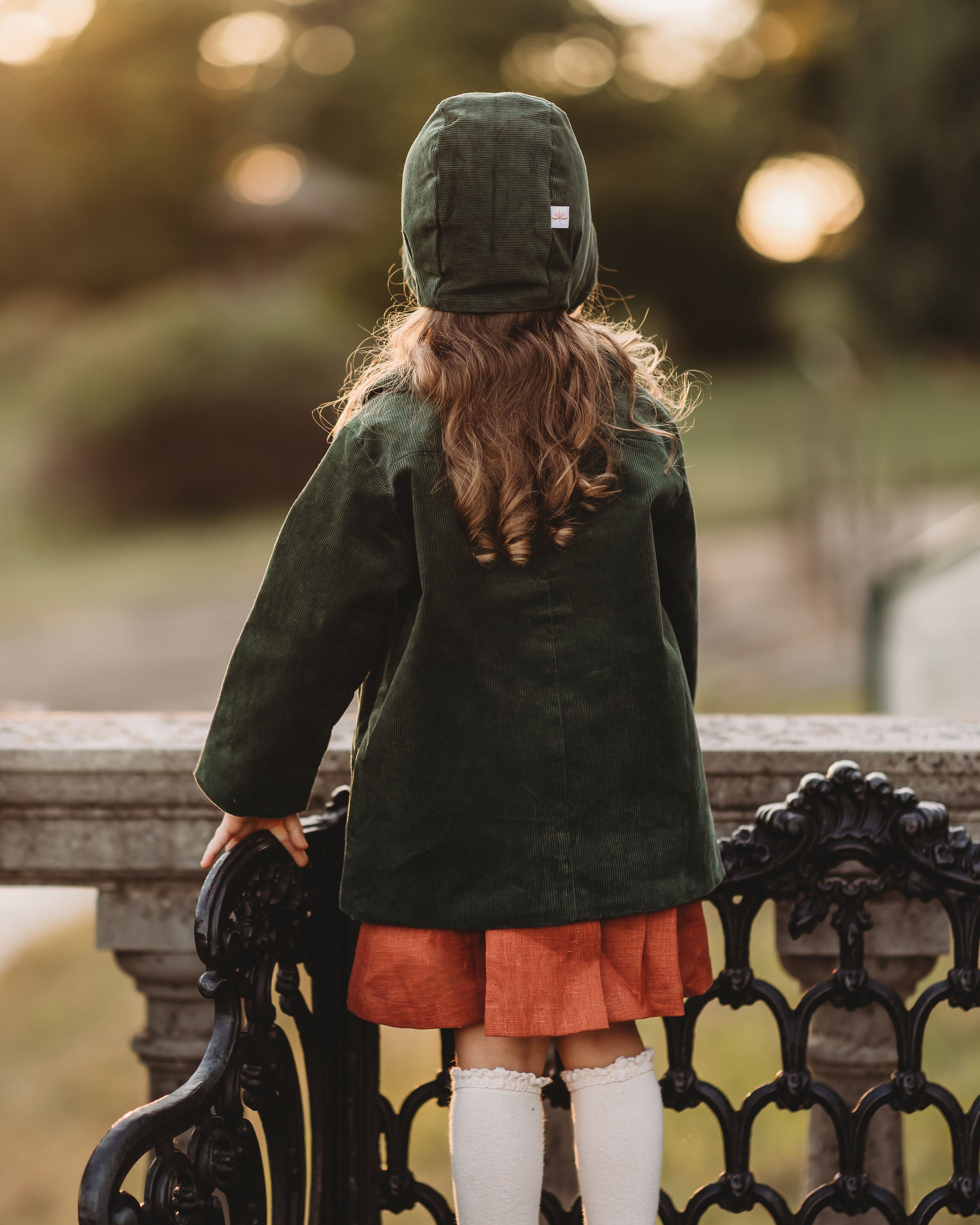 Person wearing a green coat and hat, standing on a bench with a blurred natural background