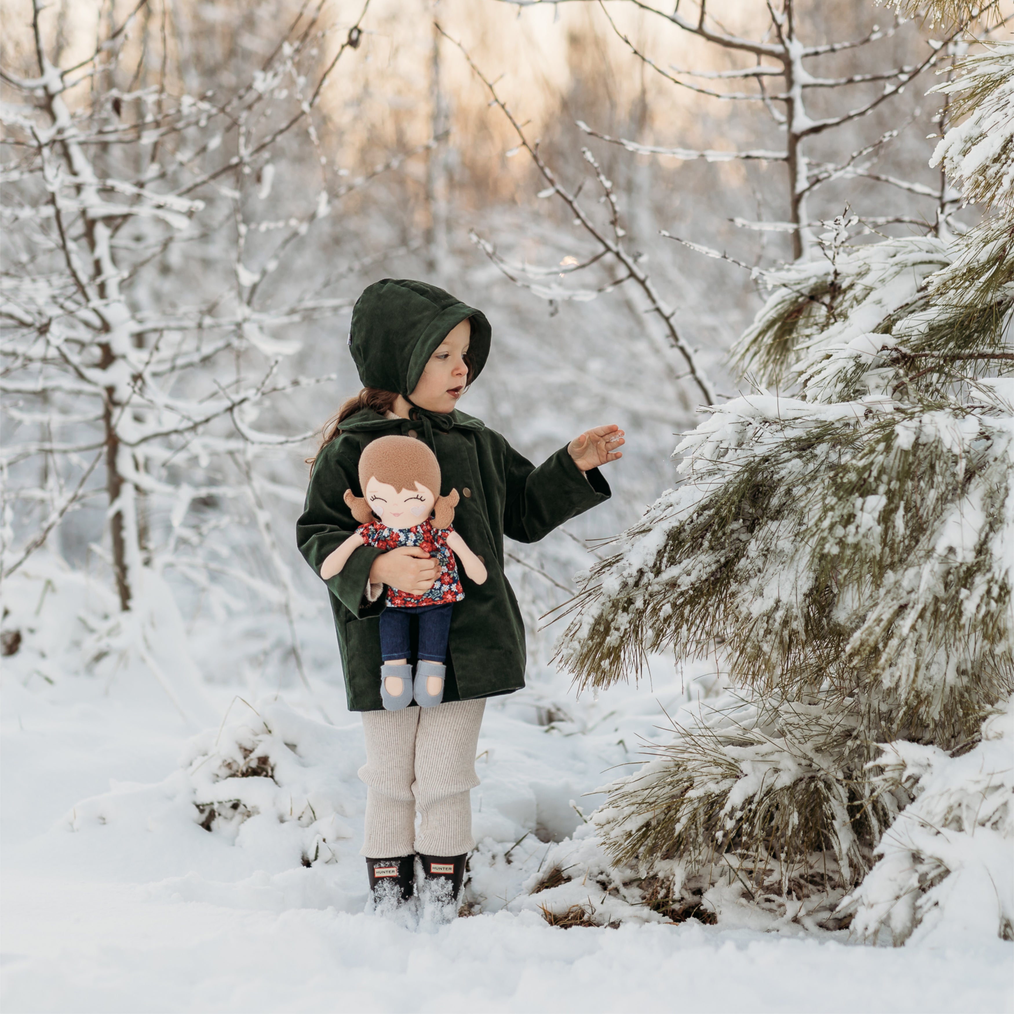 Adorable girl holding her doll next to a snow-covered tree, showcasing the Evergreen bonnet and its perfect pairing with sustainable children’s clothing.