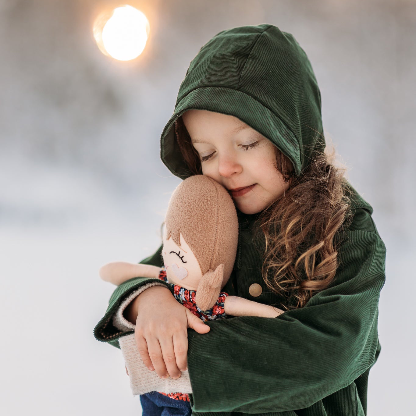 Model girl cuddling a doll in a snowy landscape, wearing the Evergreen bonnet, representing warmth and timeless children’s wear.