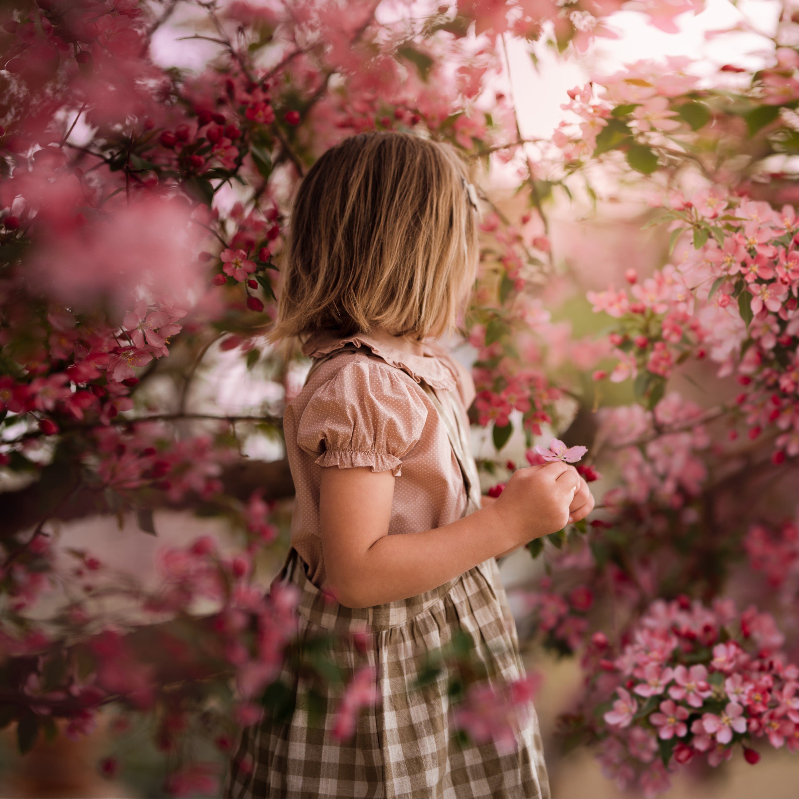 Side details of a young girl wearing a beige blouse with white Swiss dots and a matching beige gingham pinafore. The blouse features a Peter Pan collar with ruffles and short puffy sleeves, while the pinafore showcases a classic gingham pattern, perfect for playful outfits