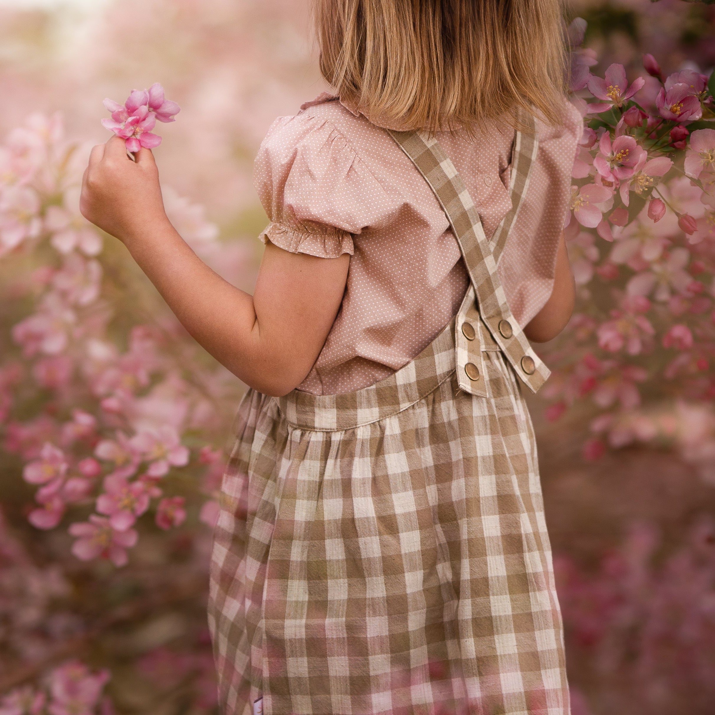 Back details of a young girl wearing a beige blouse with white Swiss dots and a matching beige gingham pinafore. The blouse features a Peter Pan collar with ruffles and short puffy sleeves, while the pinafore showcases a classic gingham pattern and adjustable straps, perfect for playful outfits.