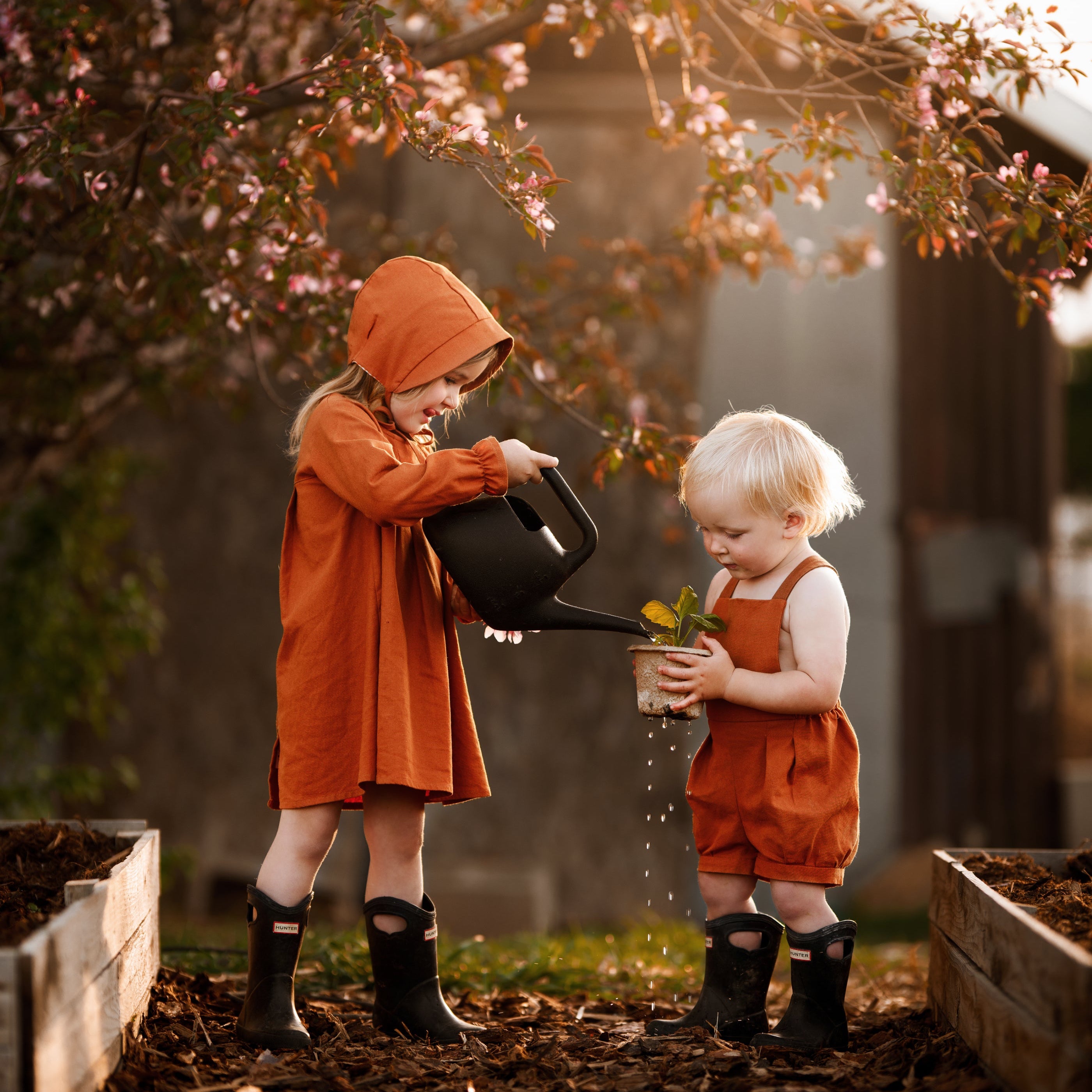 Two children in orange outfits and rain boots playing with a watering can and plant in a garden.