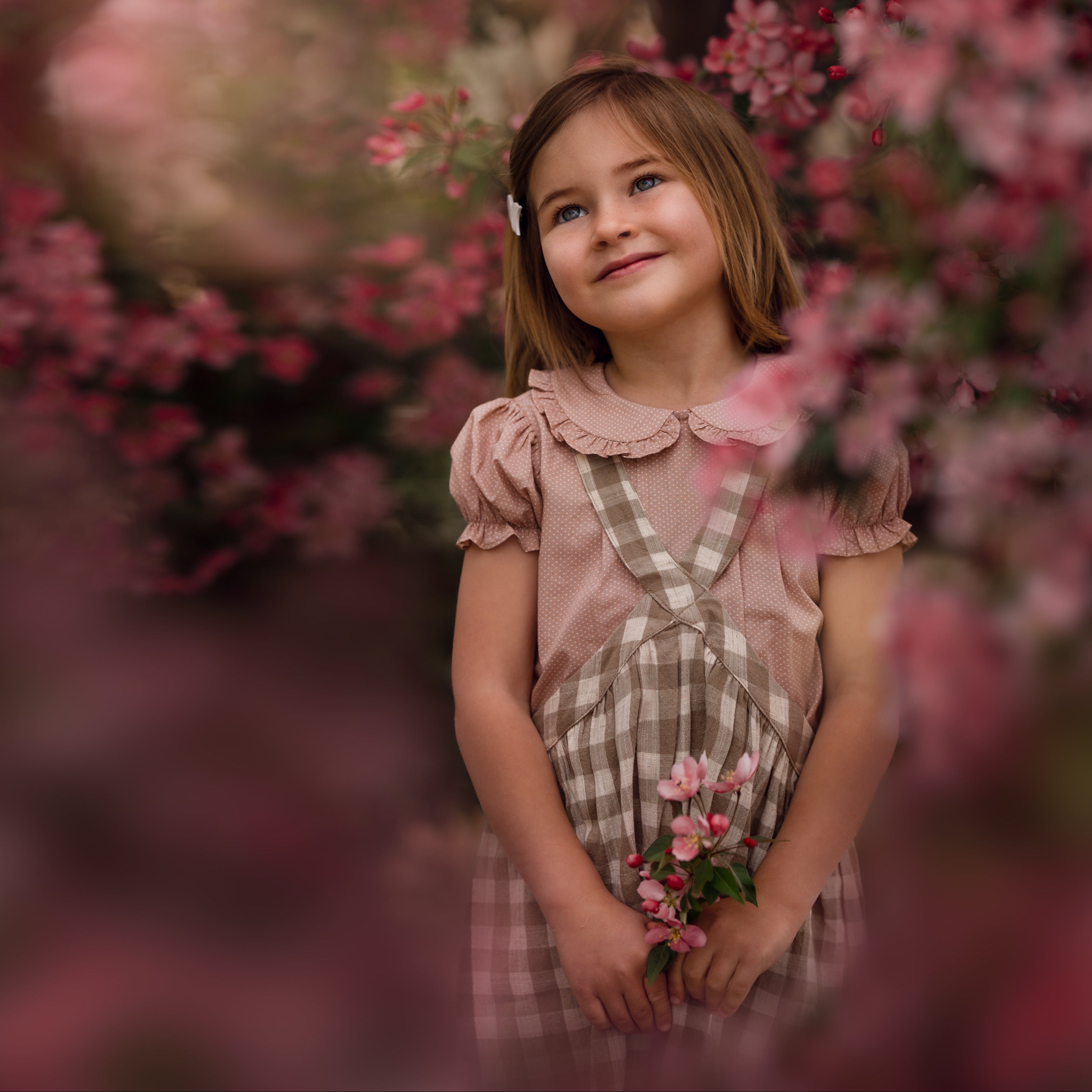Full view of a young girl wearing a beige blouse with white Swiss dots and a matching beige gingham pinafore. The blouse features a Peter Pan collar with ruffles and short puffy sleeves, while the pinafore showcases a classic gingham pattern, perfect for playful outfits.