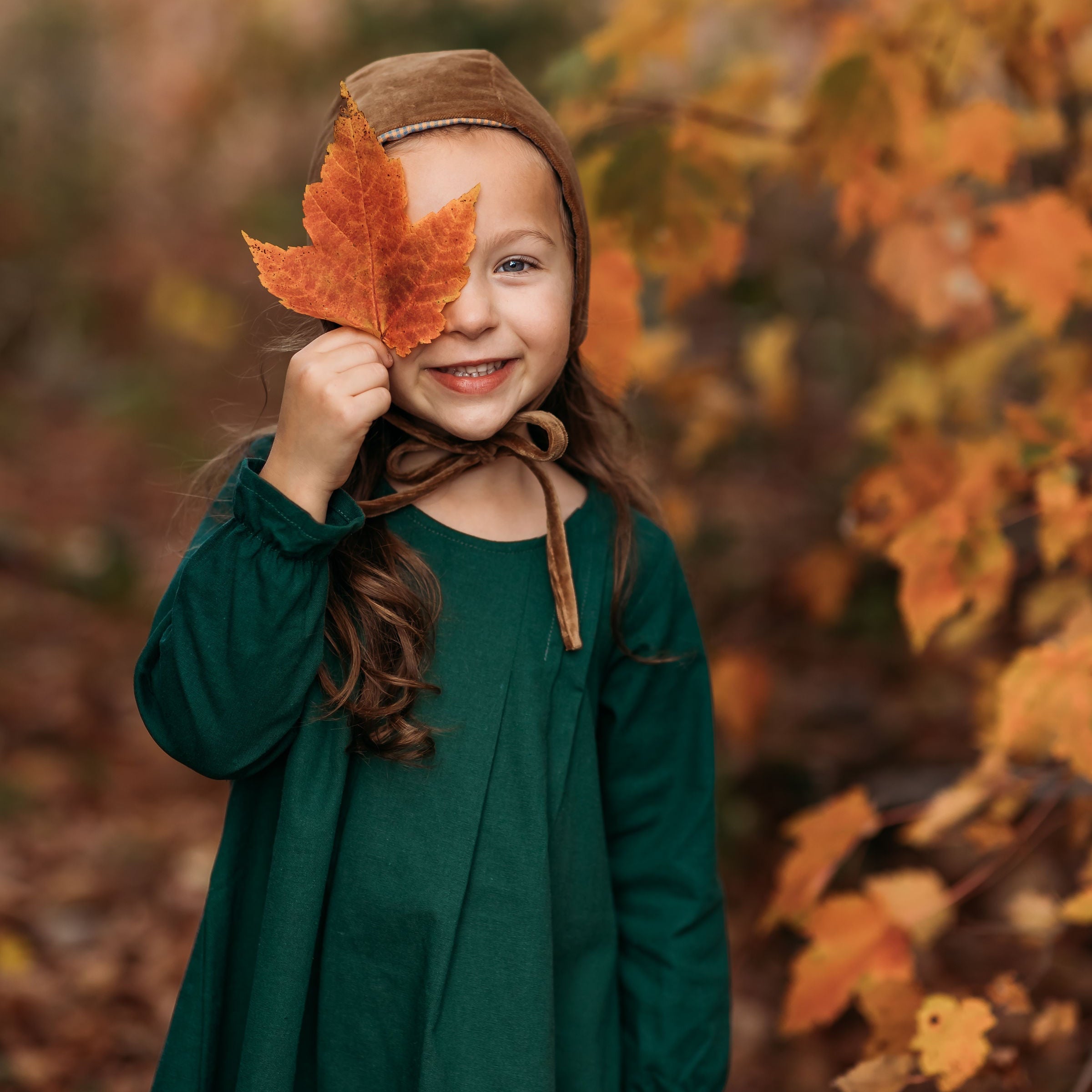 A close-up of a 4-year-old girl holding a leaf to her face while wearing the evergreen linen dress, emphasizing the delicate ruffled hem and fun details.