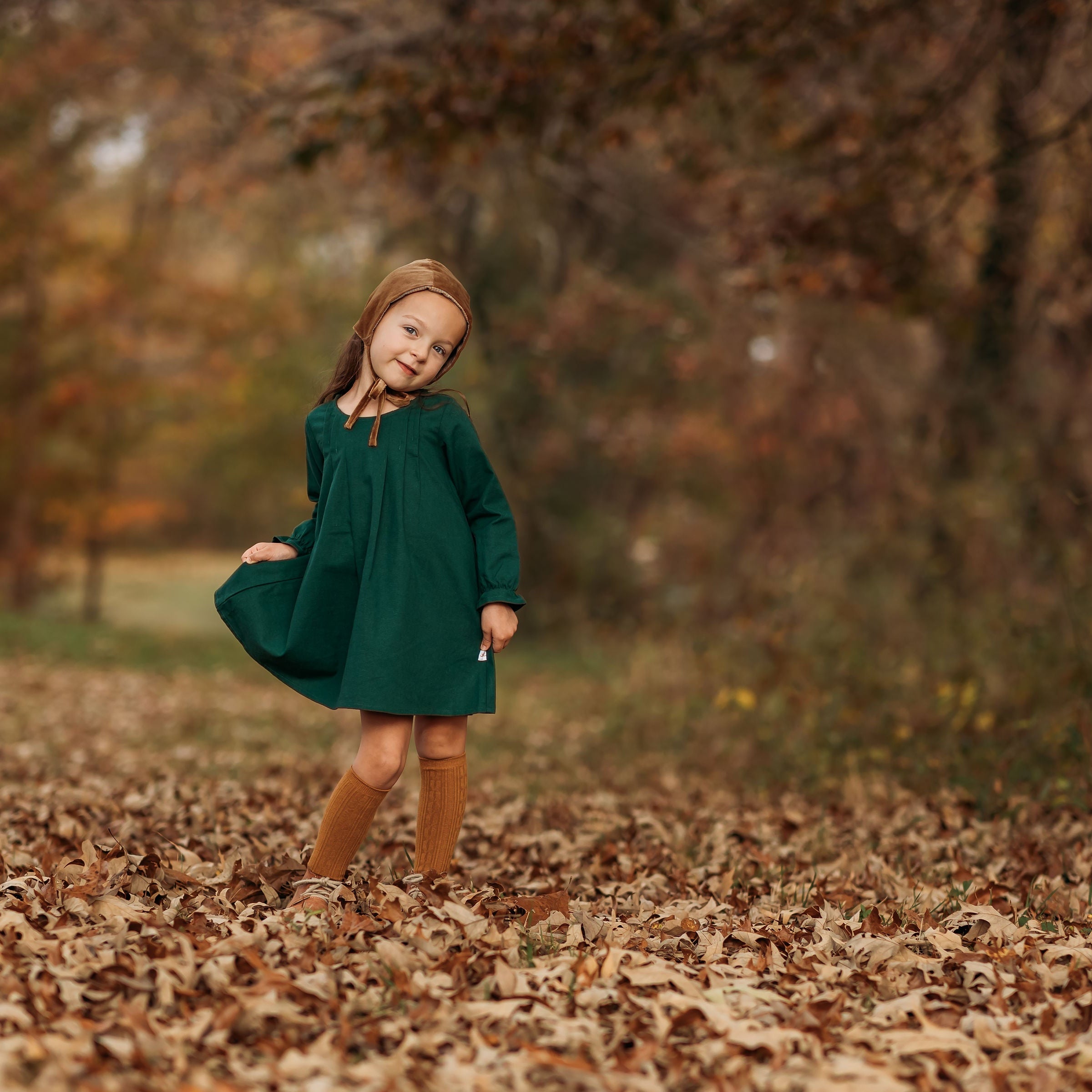 A 4-year-old girl posing sweetly while holding the tip of her dress’s skirt, showcasing the stylish fit and comfortable design of the evergreen linen dress.