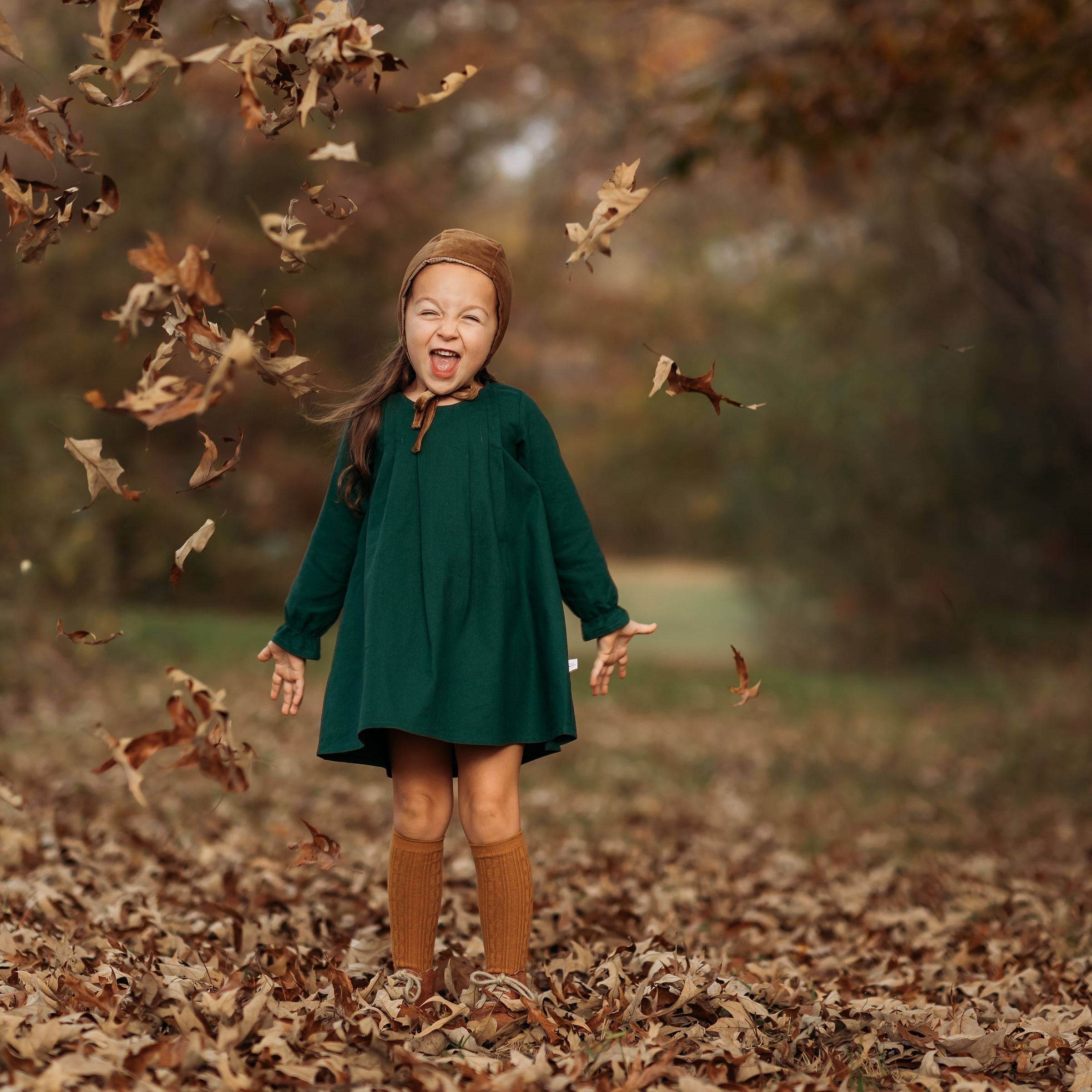 A 4-year-old girl surrounded by colorful autumn leaves, laughing joyfully in her evergreen linen dress, highlighting the playful spirit and flattering A-line shape.