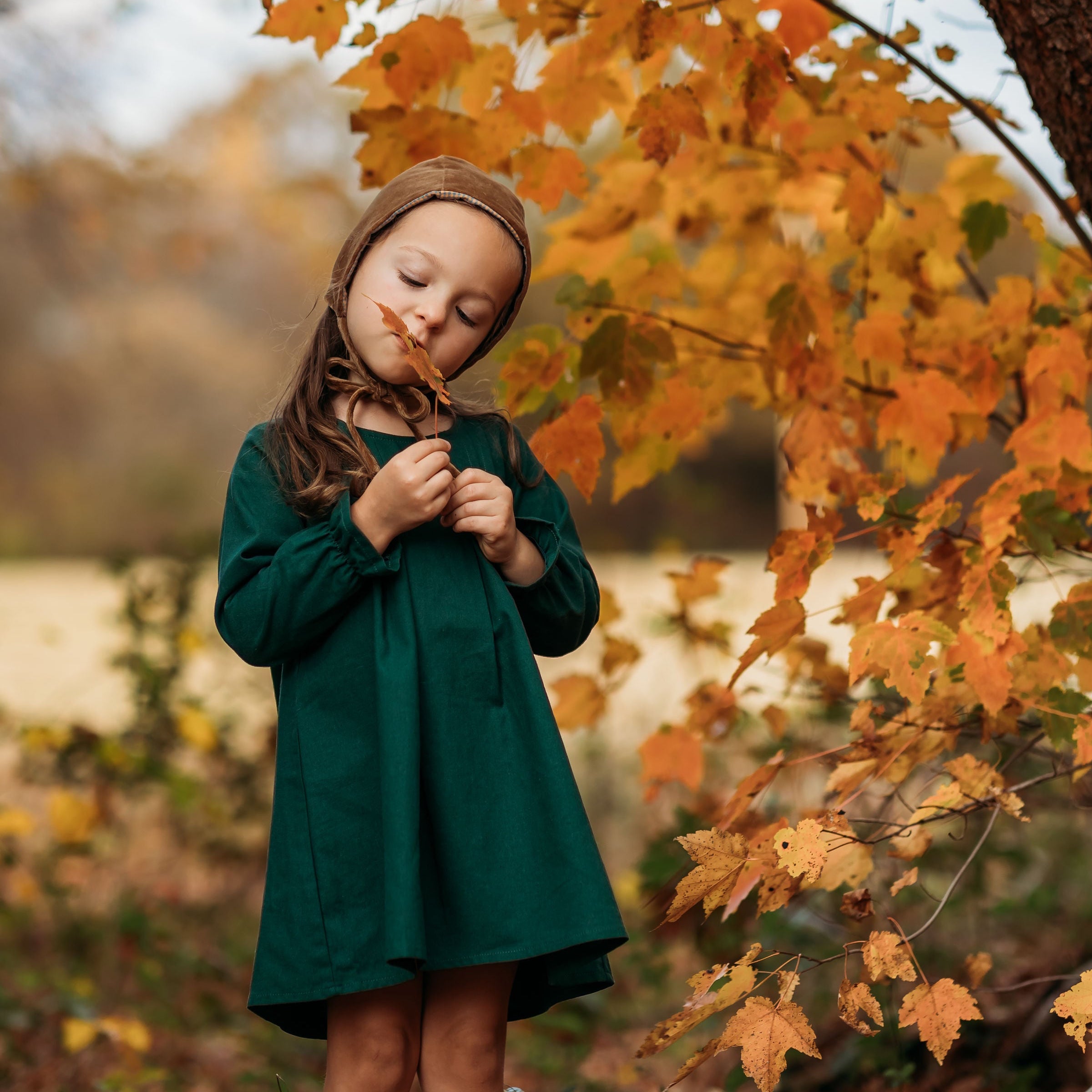 A 4-year-old girl standing in a fall setting, wearing the evergreen linen dress while holding a brown leaf, capturing the seasonal charm and style of the outfit.