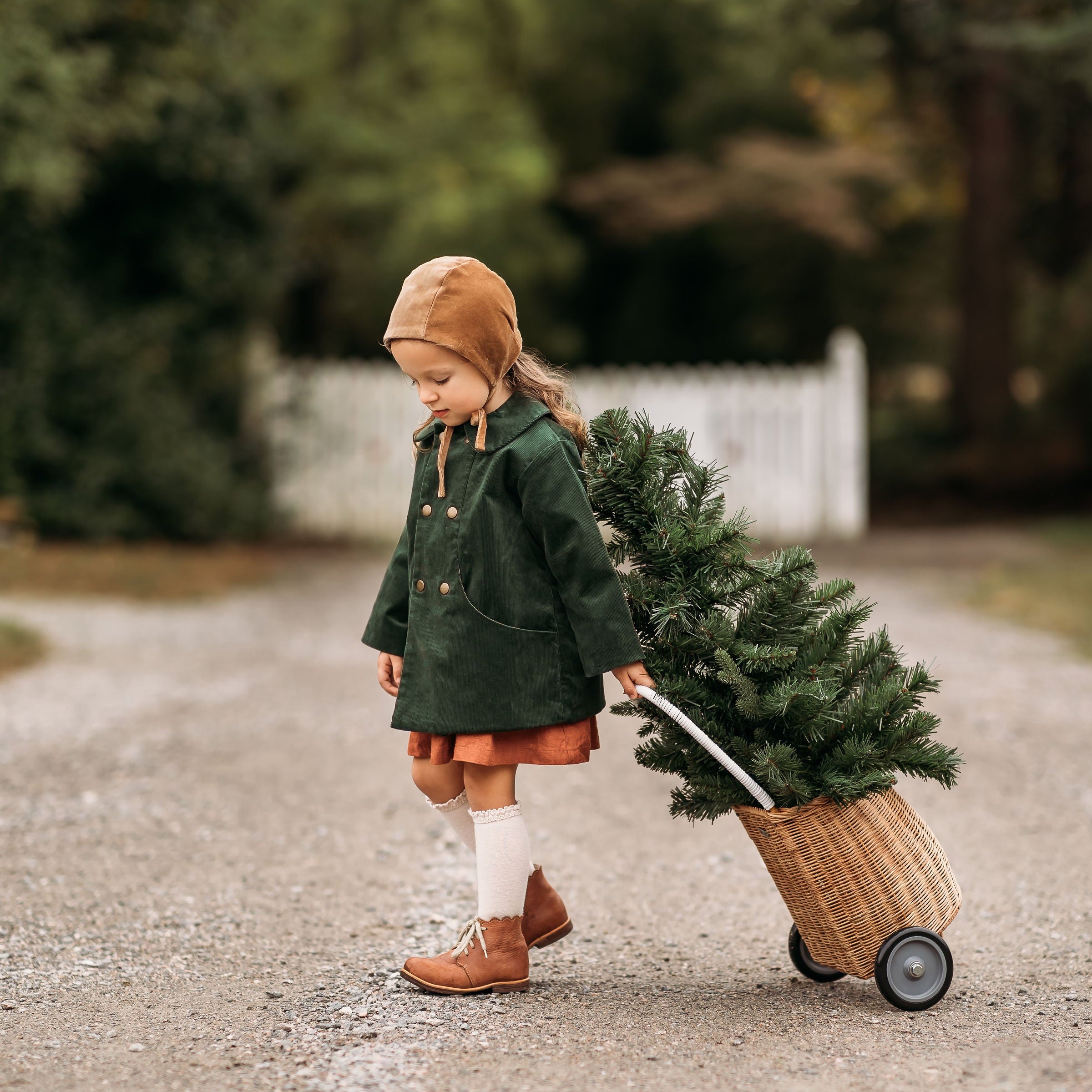 Side view of a girl in the Gold Velvet Bonnet and Evergreen Corduroy Coat, walking away with a Christmas tree, illustrating the bonnet’s elegant style and practicality for special celebrations.