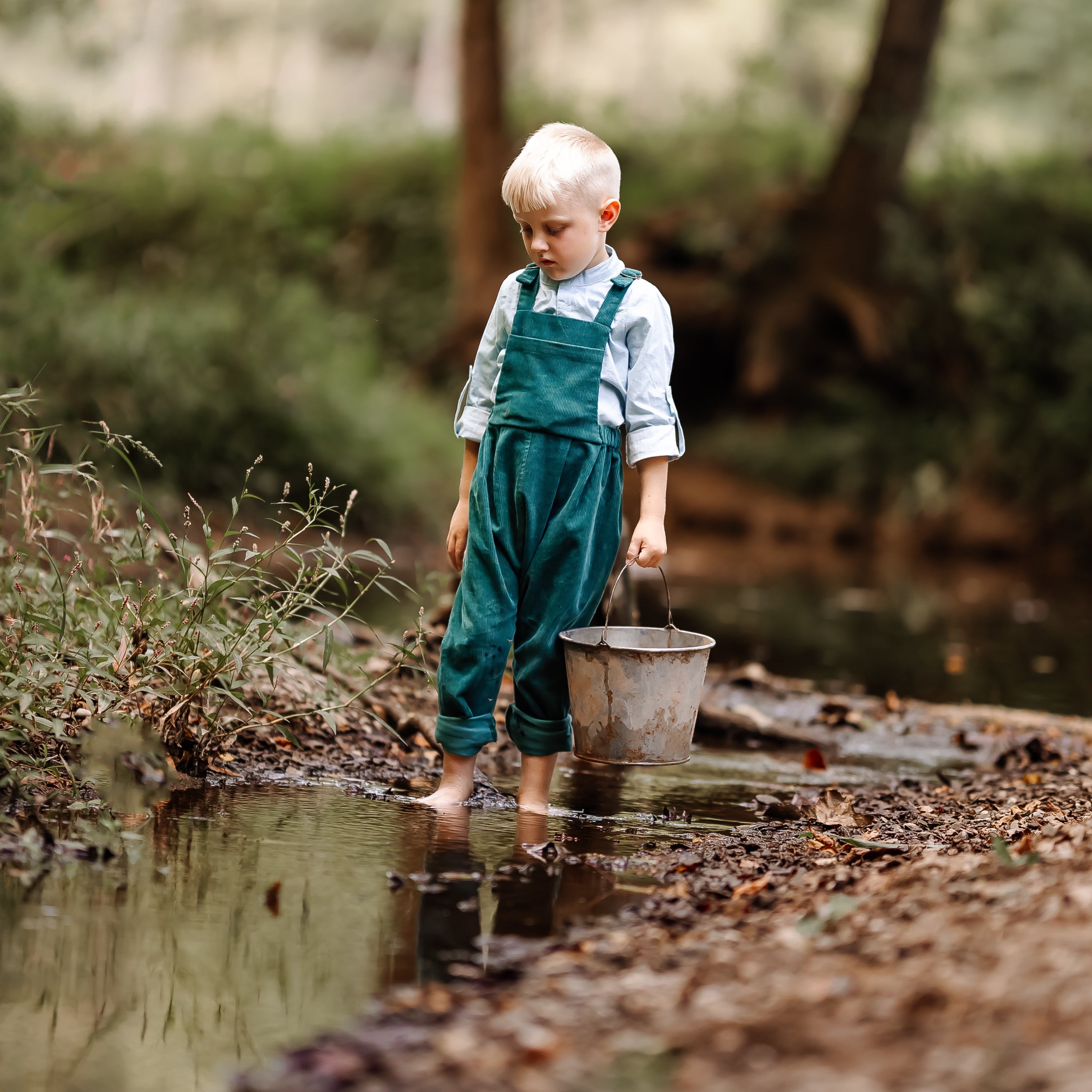 Child in green overalls holding a bucket by a stream in a forest