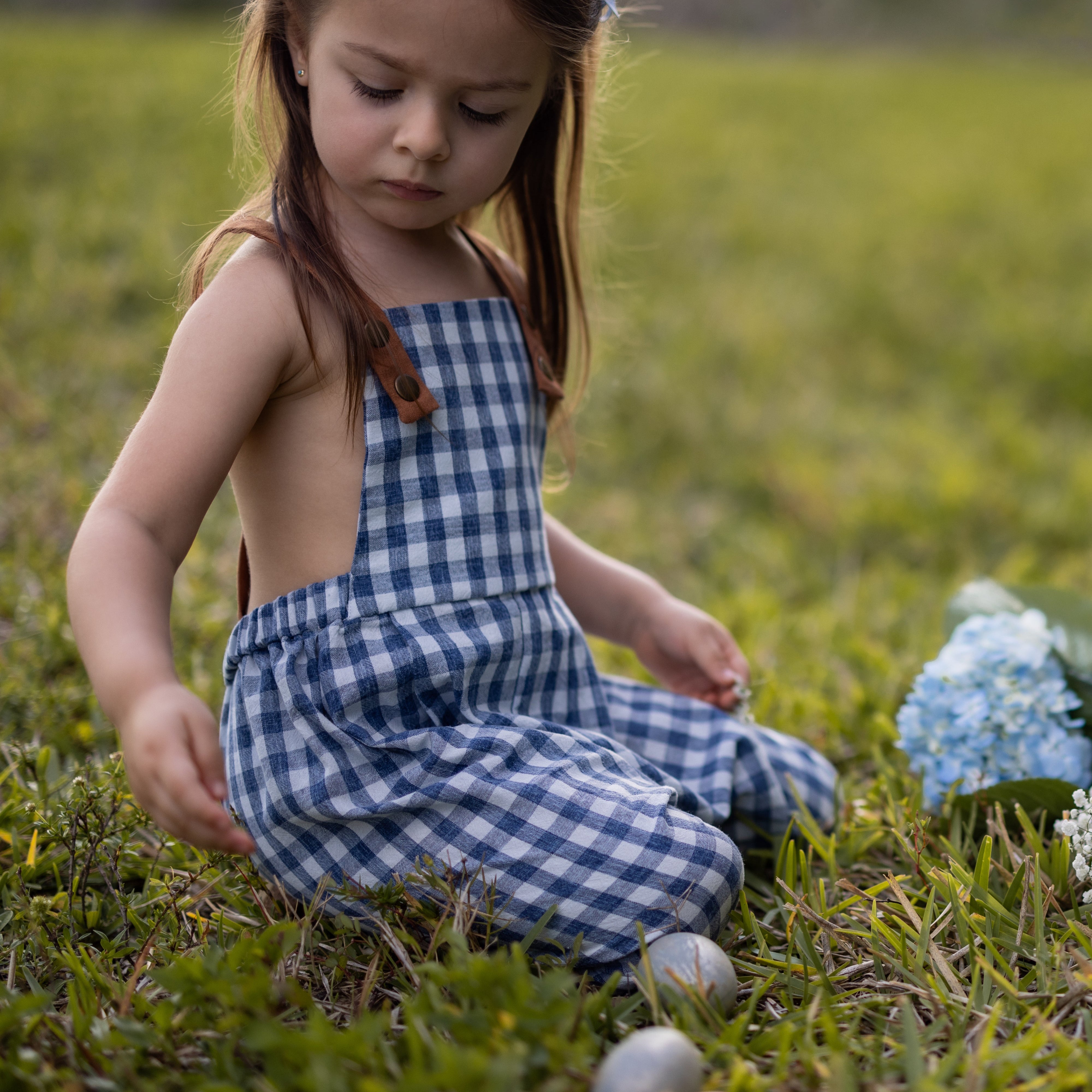 A little girl sitting down and playing while wearing Blue Gingham Linen Long Overalls, enjoying her Easter festivities. The gender-neutral design showcases her playful spirit, while the breathable fabric ensures comfort during her activities. Stylish pleats enhance the overall look, and the durable leather-looking straps along with an adjustable fit make these overalls perfect for active little ones, all by a brand dedicated to sustainable fashion.