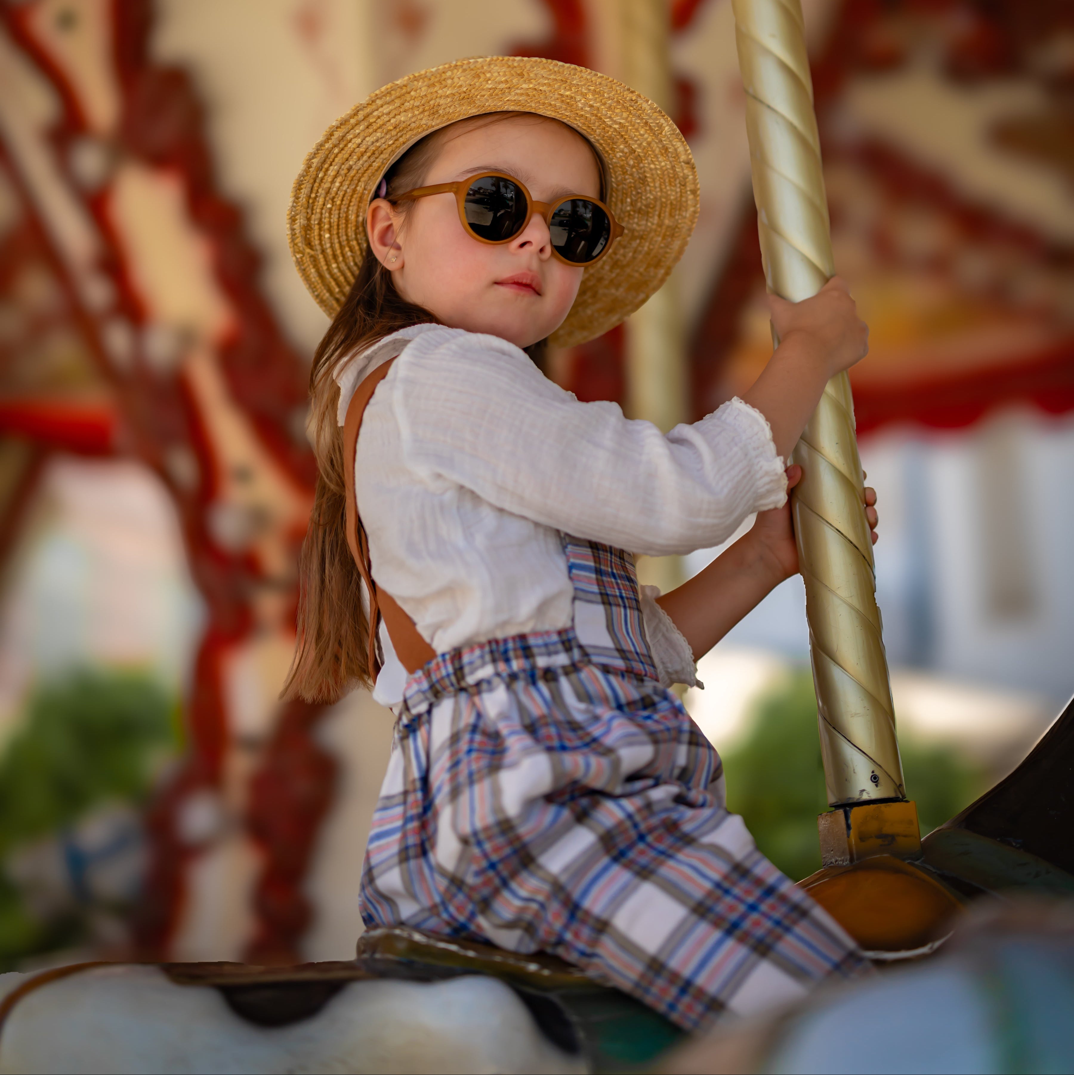 Child on a carousel holding onto a pole, wearing a straw hat and sunglasses and plaid linen overalls.
