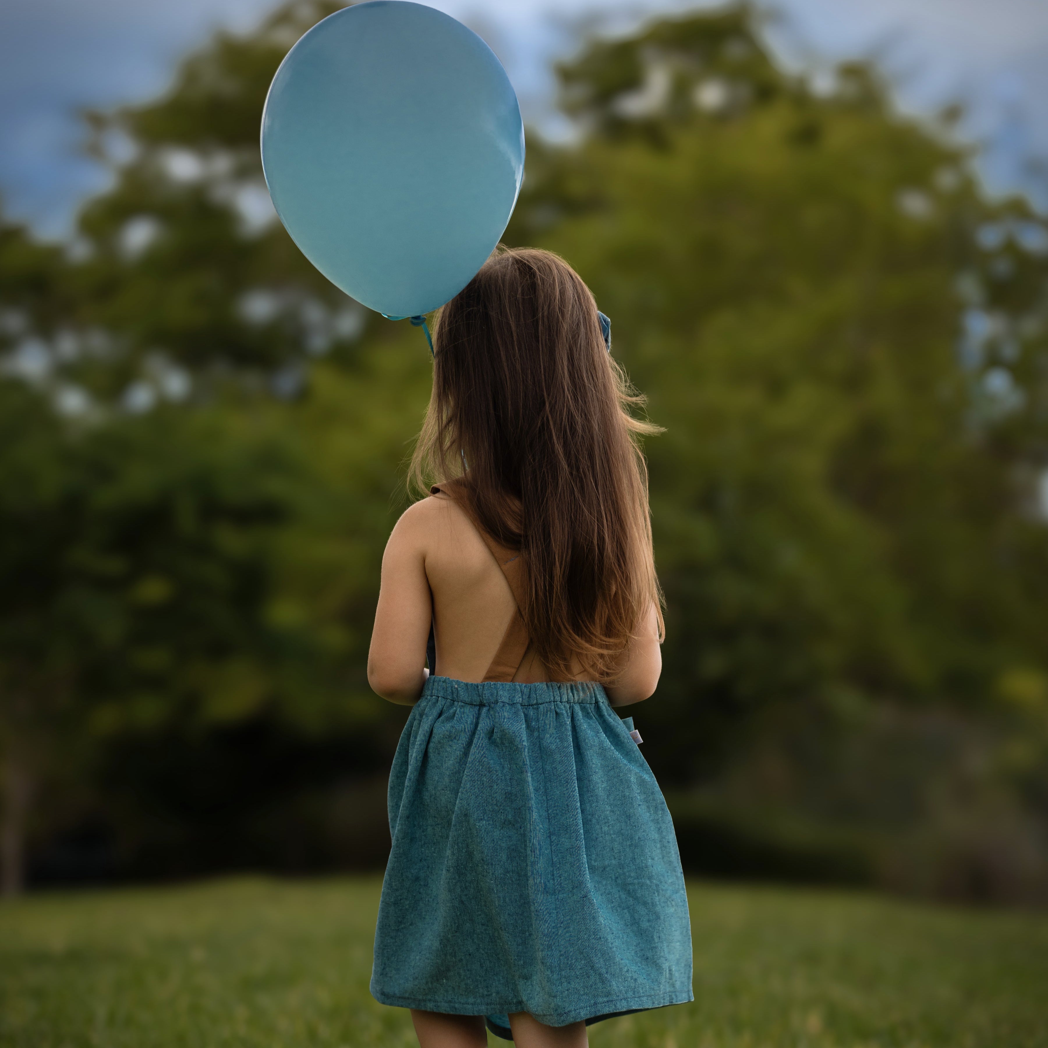 A little girl model walking away in the Blue Jay pinafore, showcasing the back in a summer setting - Handmade from repurposed linen in our NYC studio, this classic design features durable leather-looking straps and adjustable lengths for a perfect fit. The generous pocket is great for storing little treasures, making it a stylish choice for summer adventures.