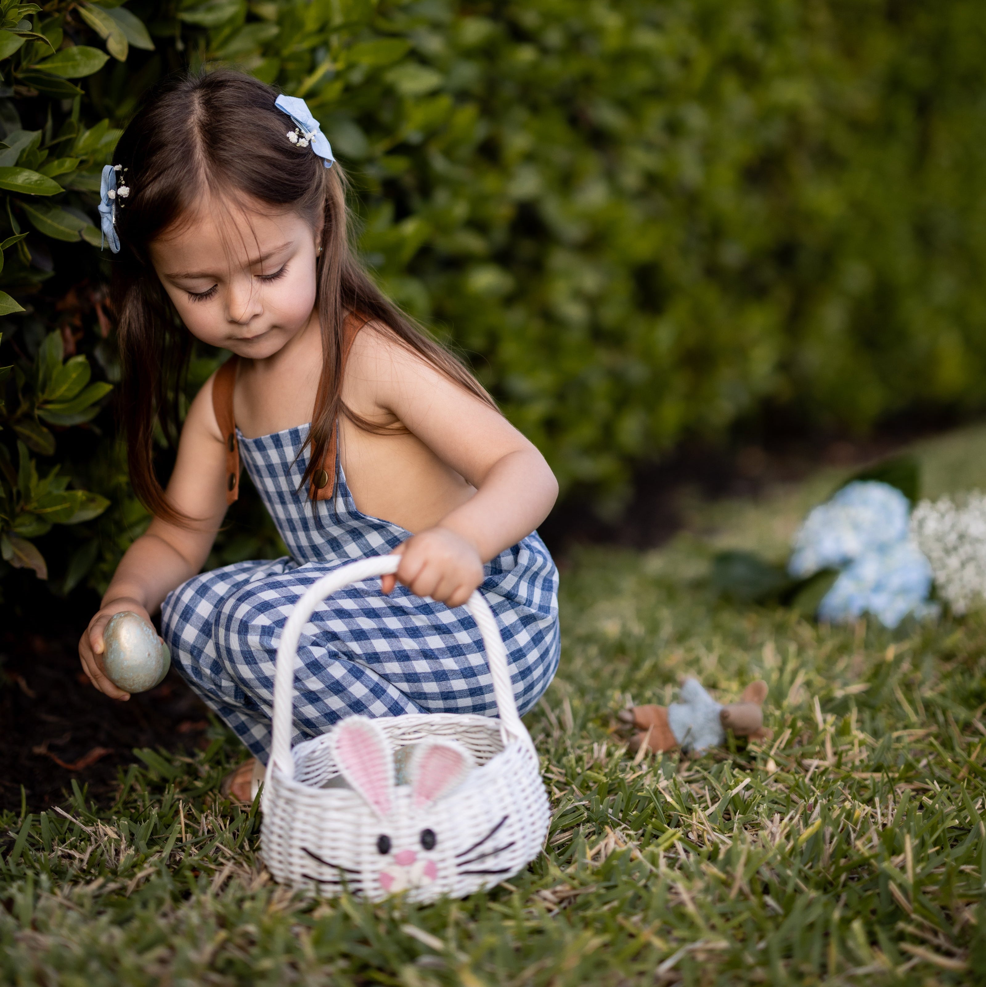 A little girl picking Easter eggs while wearing Blue Gingham Linen Long Overalls, fully immersed in the joy of the holiday. The playful, gender-neutral design allows her to move freely as she gathers her treasures, and the breathable fabric keeps her comfortable during the fun. Stylish pleats add a charming touch, while durable leather-looking straps and an adjustable fit make these overalls a practical choice for her adventurous spirit, all from a brand committed to sustainable fashion for children.