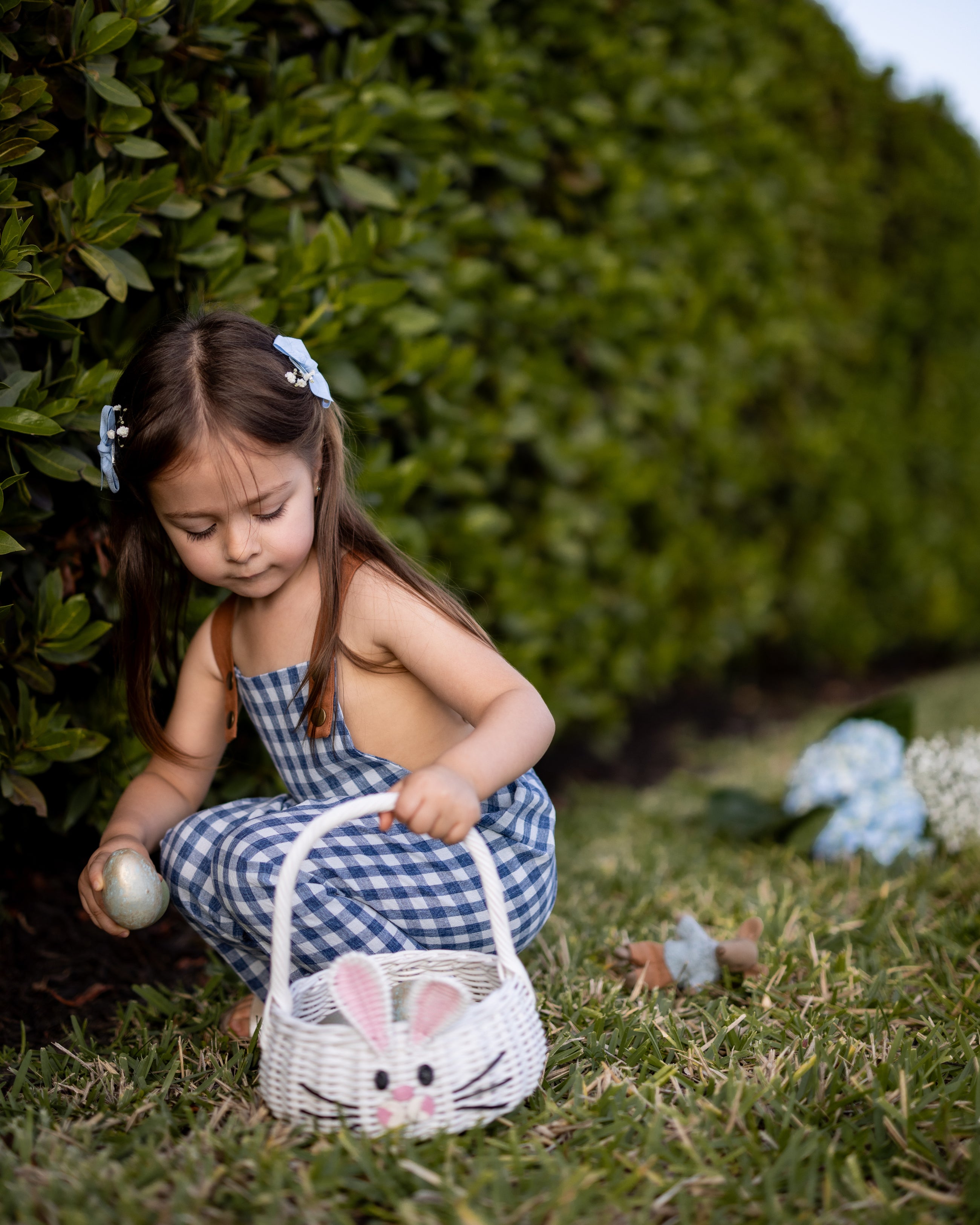 A little girl picking Easter eggs while wearing Blue Gingham Linen Long Overalls, fully immersed in the joy of the holiday. The playful, gender-neutral design allows her to move freely as she gathers her treasures, and the breathable fabric keeps her comfortable during the fun. Stylish pleats add a charming touch, while durable leather-looking straps and an adjustable fit make these overalls a practical choice for her adventurous spirit, all from a brand committed to sustainable fashion for children.