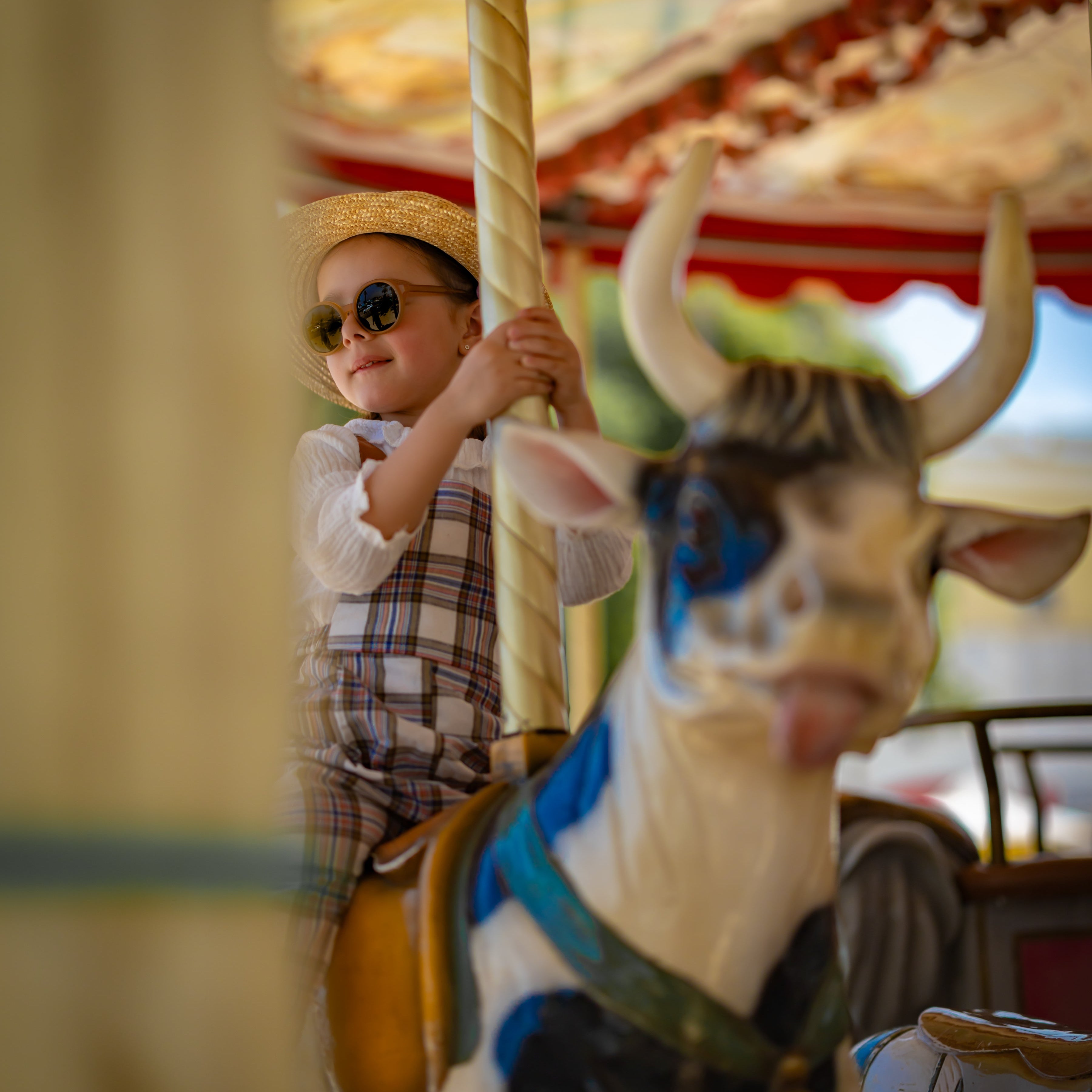 Child on a carousel with a cow figure, wearing sunglasses and a hat and plaid linen overalls.
