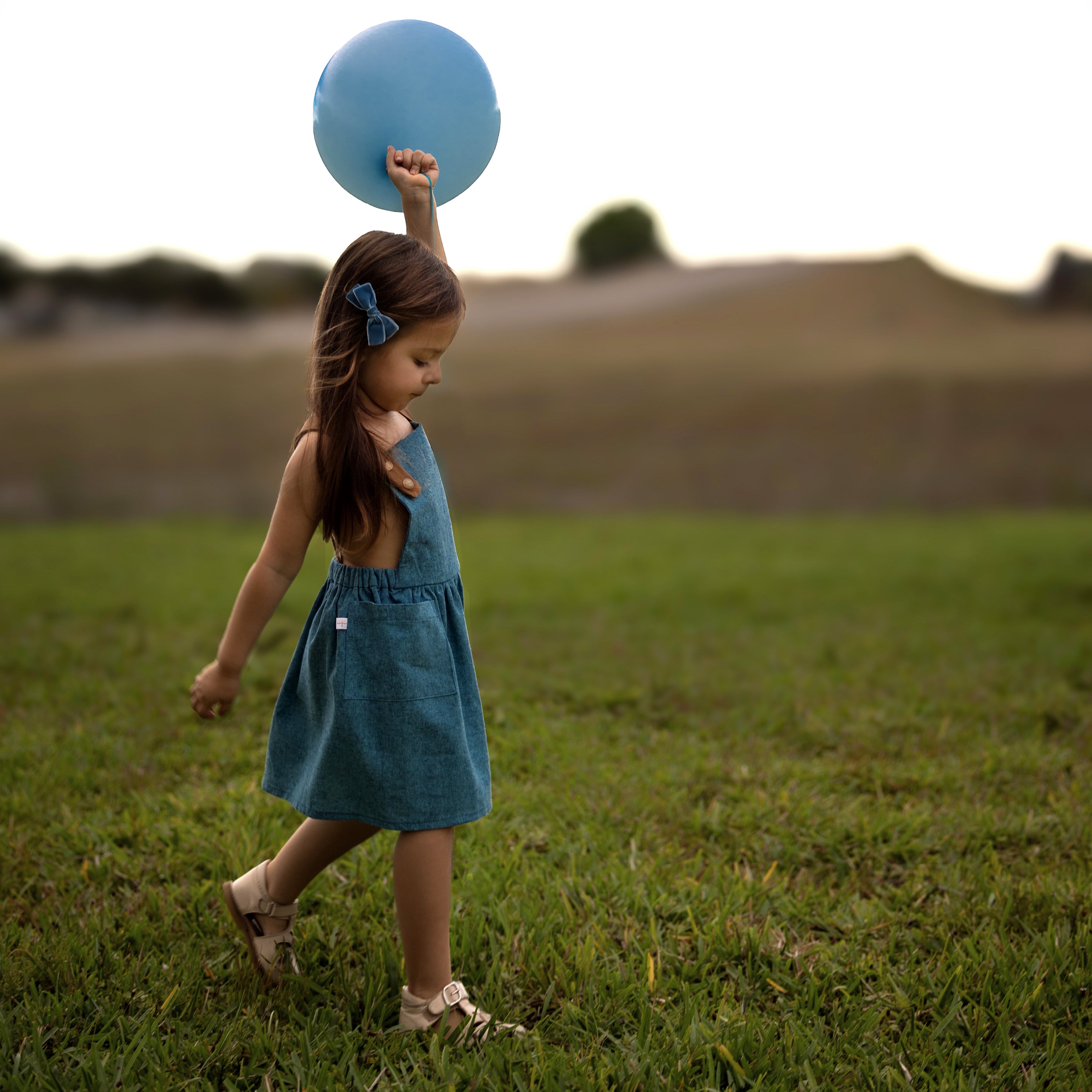 Side view of a little girl model wearing the Blue Jay pinafore while holding a blue balloon in the summer - This classic and timeless design is handmade from repurposed linen in our NYC studio. The pinafore is designed to be worn on its own, allowing your little girl to stay stylish and cool. The durable leather-looking straps add visual appeal, while the adjustable straps in two lengths ensure a perfect fit. A generous pocket is perfect for storing little treasures, making it an ideal choice for summer fun