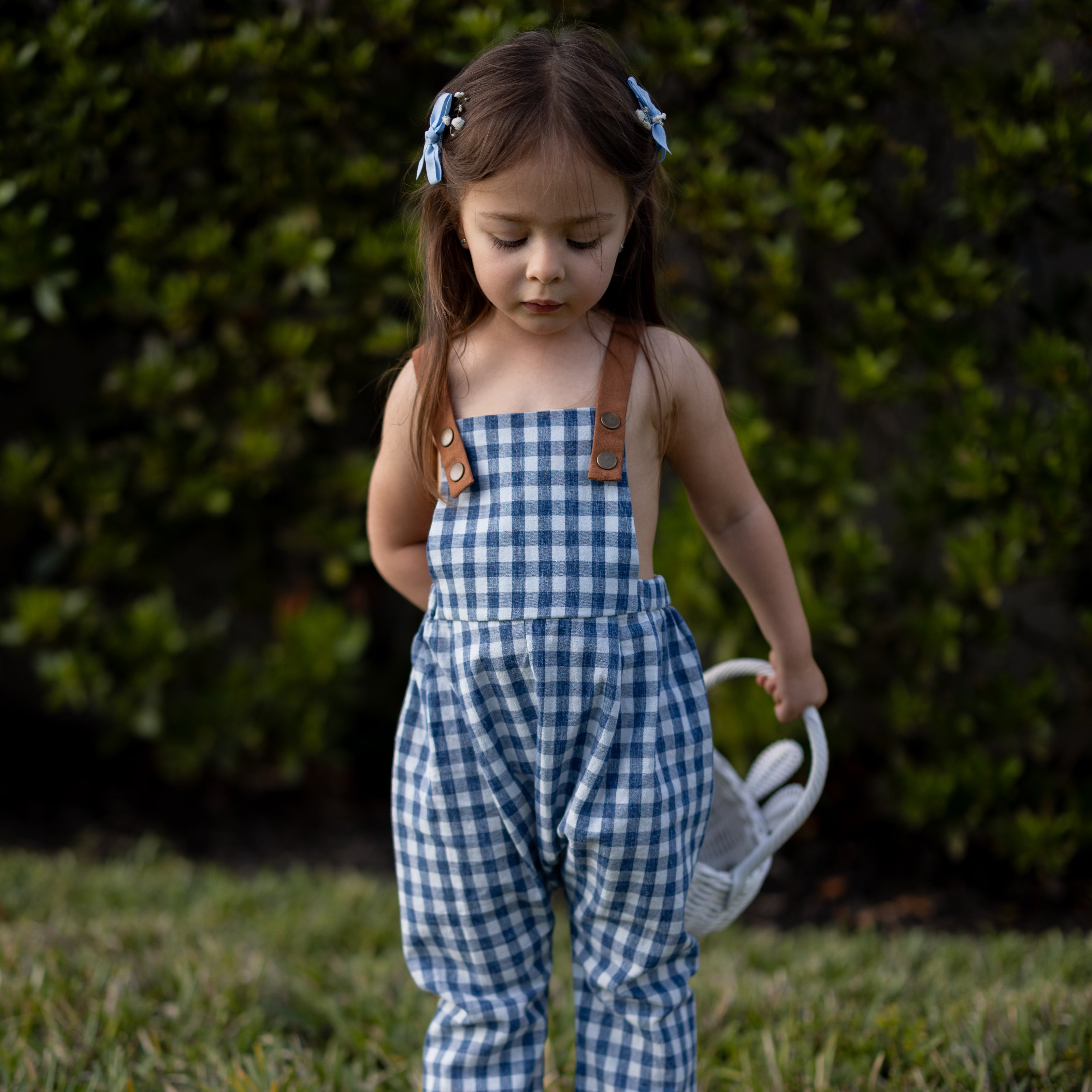 A little girl joyfully wearing Blue Gingham Linen Long Overalls, showcasing the playful and gender-neutral design perfect for Easter celebrations. The breathable fabric keeps her comfortable, while stylish pleats add a charming touch. The durable leather-looking straps and adjustable fit ensure she looks adorable and feels great during her festive activities.