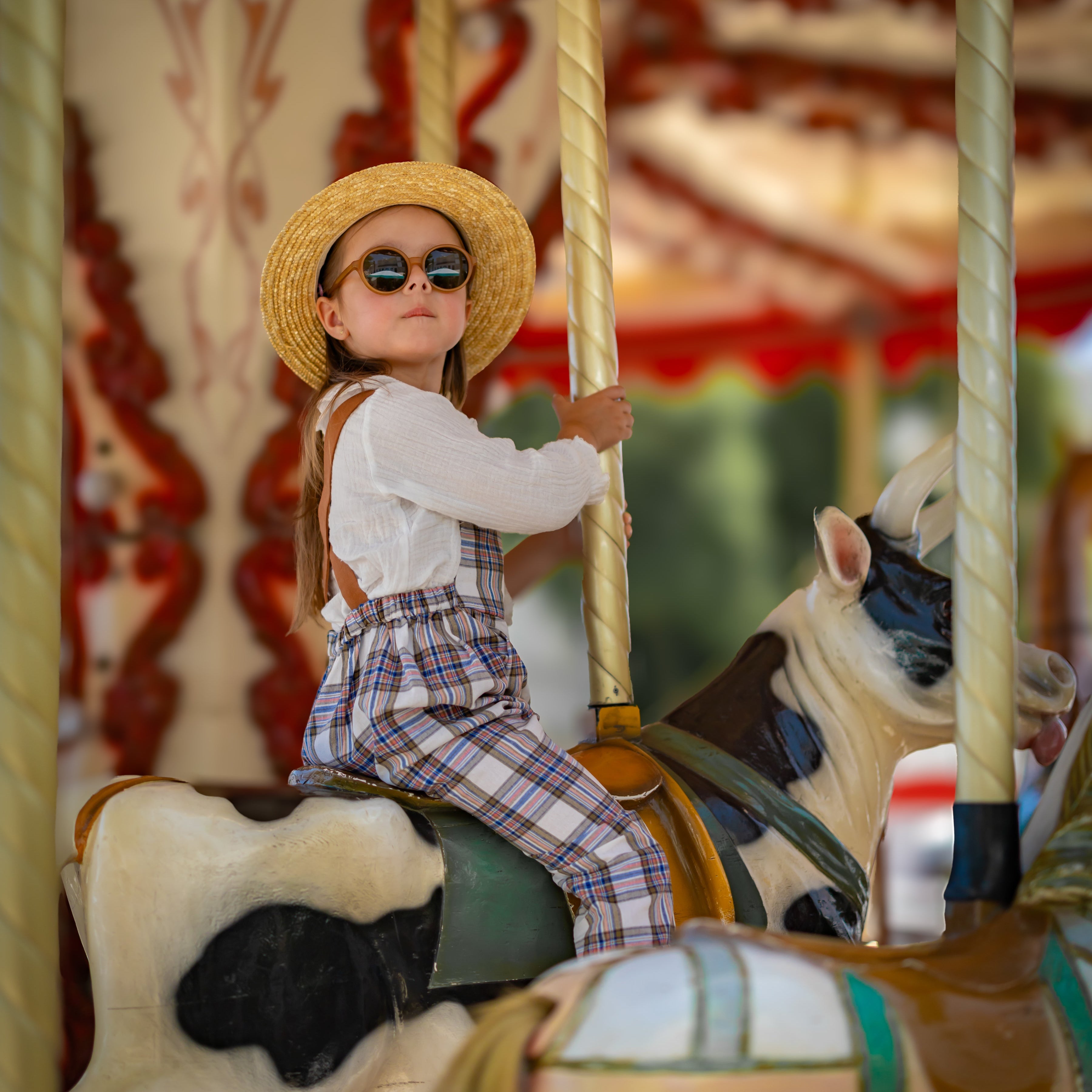 Child on a carousel with a horse, wearing a straw hat and sunglasses and plaid linen overalls.