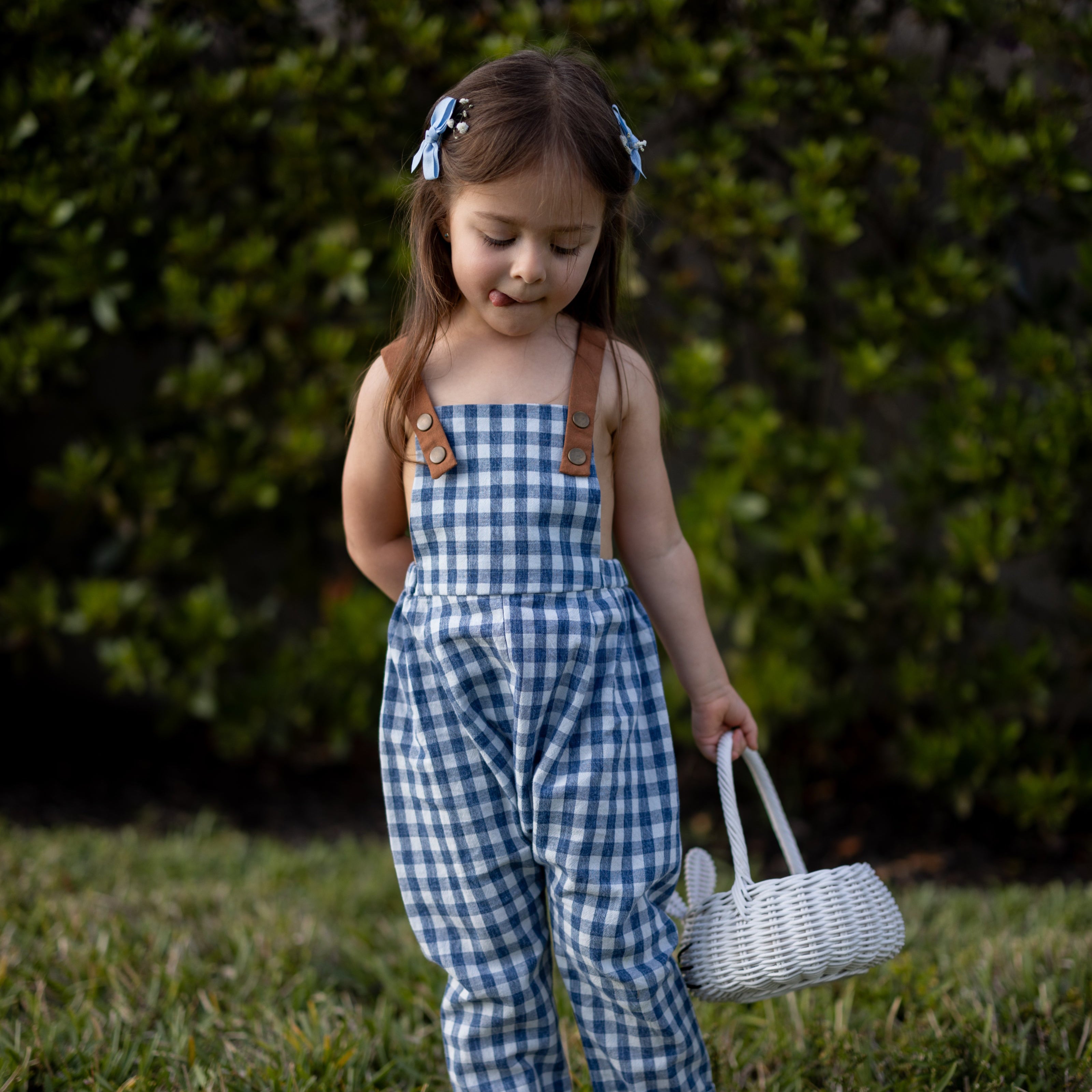 A little girl playfully sticking her tongue out while wearing Blue Gingham Linen Long Overalls, capturing a joyful moment during Easter. The gender-neutral design and breathable fabric allow for carefree movement, while the stylish pleats add a touch of elegance. Durable leather-looking straps and an adjustable fit make these overalls both fun and practical, reflecting the brand's commitment to sustainable fashion for children.