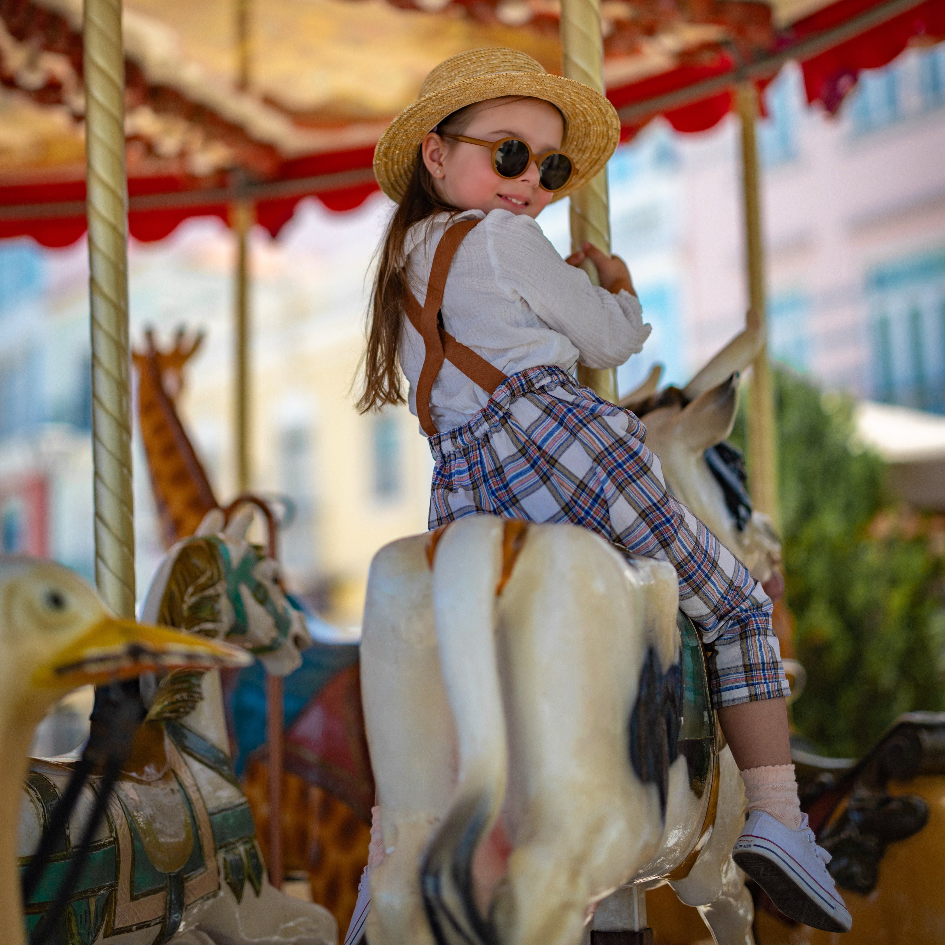 Child on a carousel with colorful background wearing a hat and plaid linen overalls.